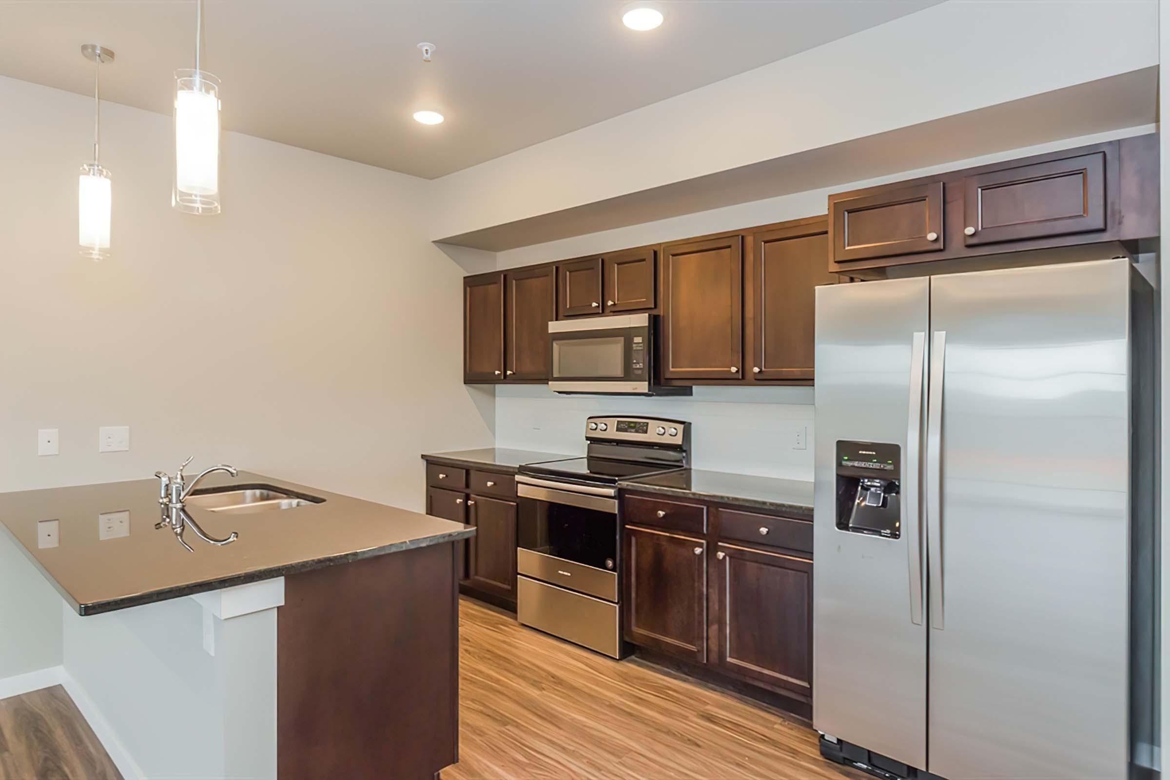 Modern kitchen featuring dark wooden cabinetry, stainless steel appliances including a refrigerator and stove, a sleek countertop with a sink, and overhead pendant lighting, all set against a neutral wall color and wooden flooring.