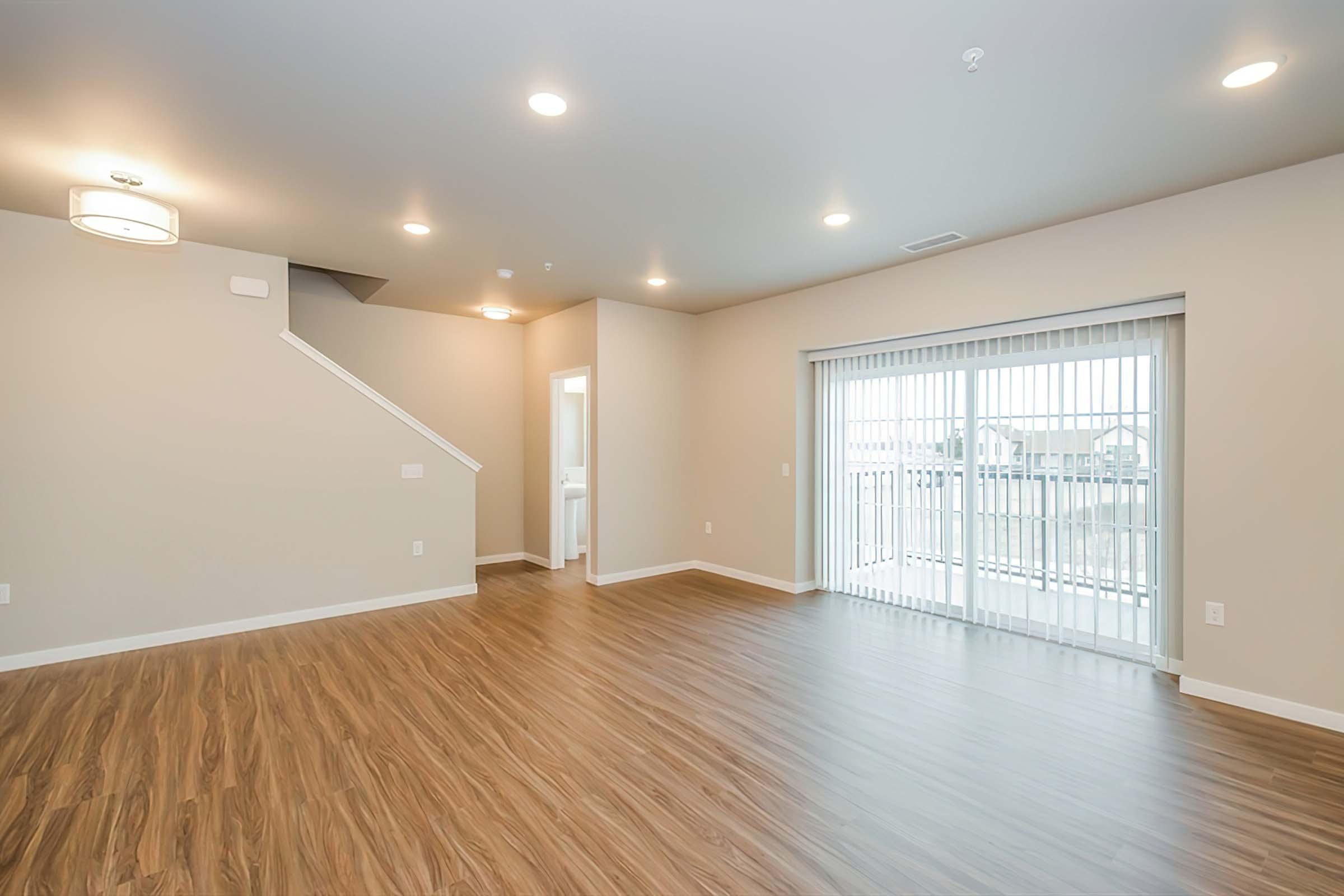Spacious interior of a modern living room with light-colored walls and hardwood flooring. Features include a staircase on the left, a door leading to another room, and sliding glass doors opening to a balcony, providing natural light. Ceiling lights illuminate the open space.