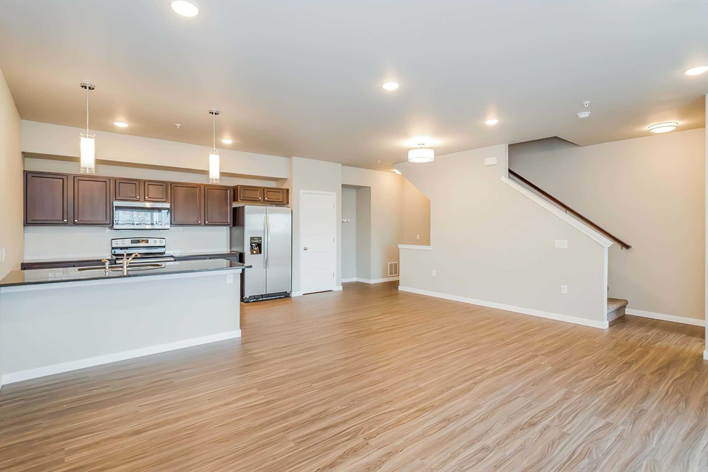 A spacious, modern living area featuring hardwood-style flooring, a kitchen with dark cabinetry and stainless steel appliances, and stylish light fixtures. The open layout includes a staircase, providing a bright and inviting atmosphere with ample natural light.
