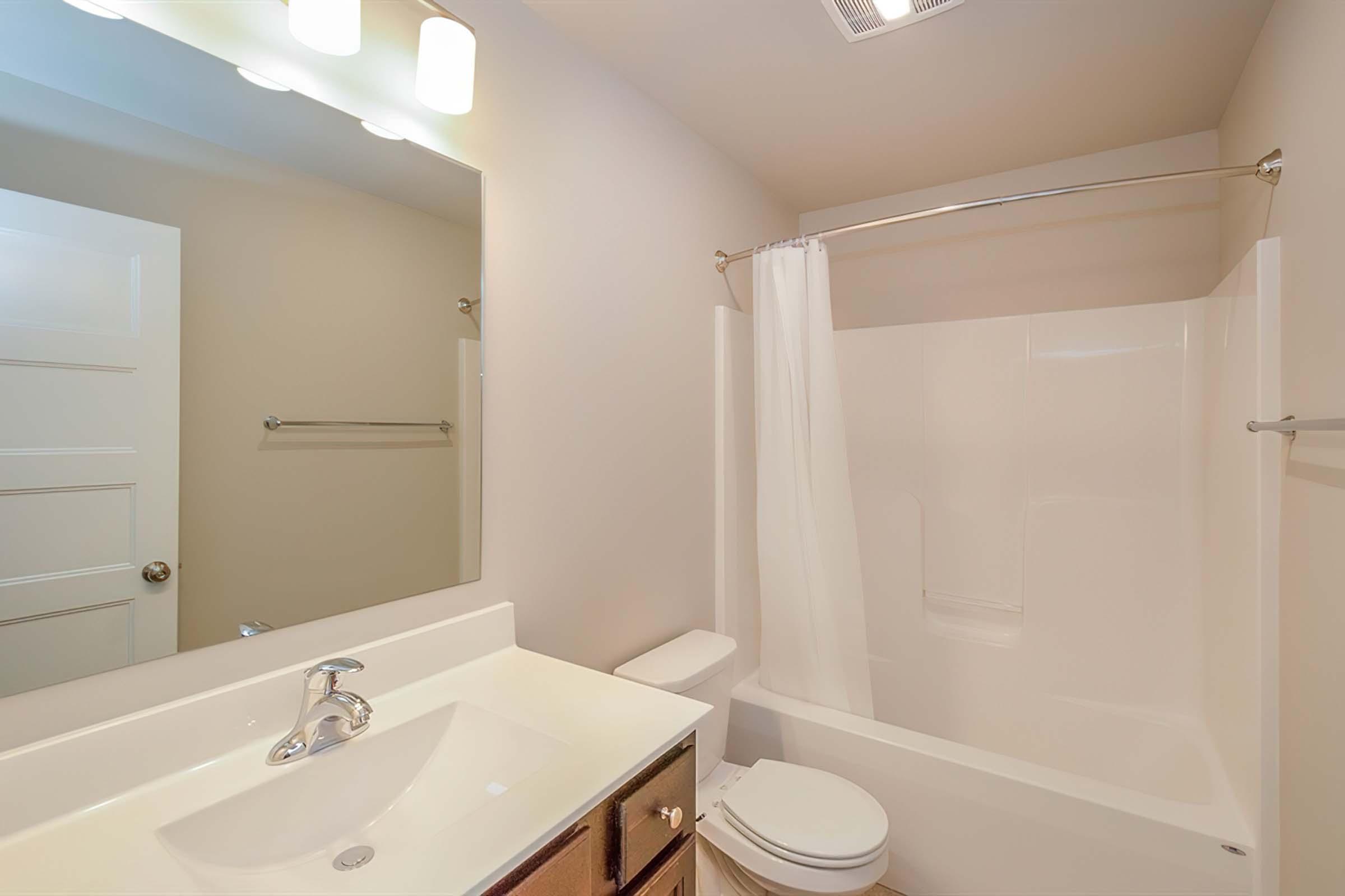A clean bathroom featuring a white bathtub with a shower curtain, a white toilet, and a countertop with a sink. The wall is light-colored, and there’s a large mirror above the sink. Two light fixtures are mounted above the mirror, creating a bright atmosphere in the space.