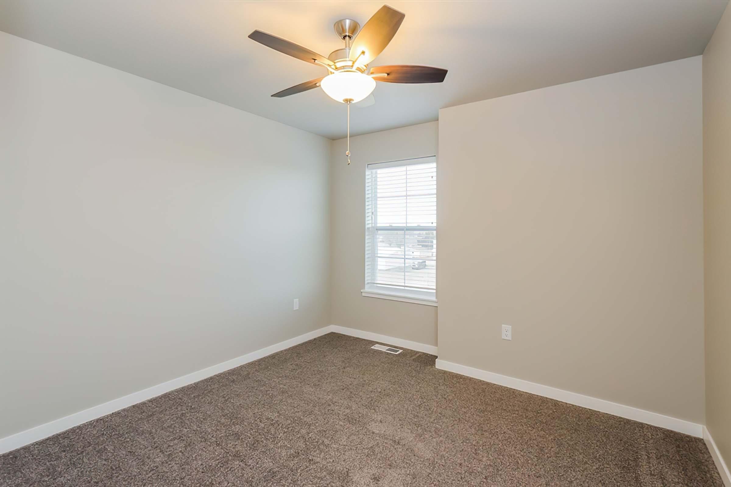 A well-lit empty room featuring light-colored walls, a ceiling fan with a light fixture, and a window with vertical blinds. The floor is covered with light brown carpet, creating a cozy atmosphere. The room is spacious and ready for furnishing.