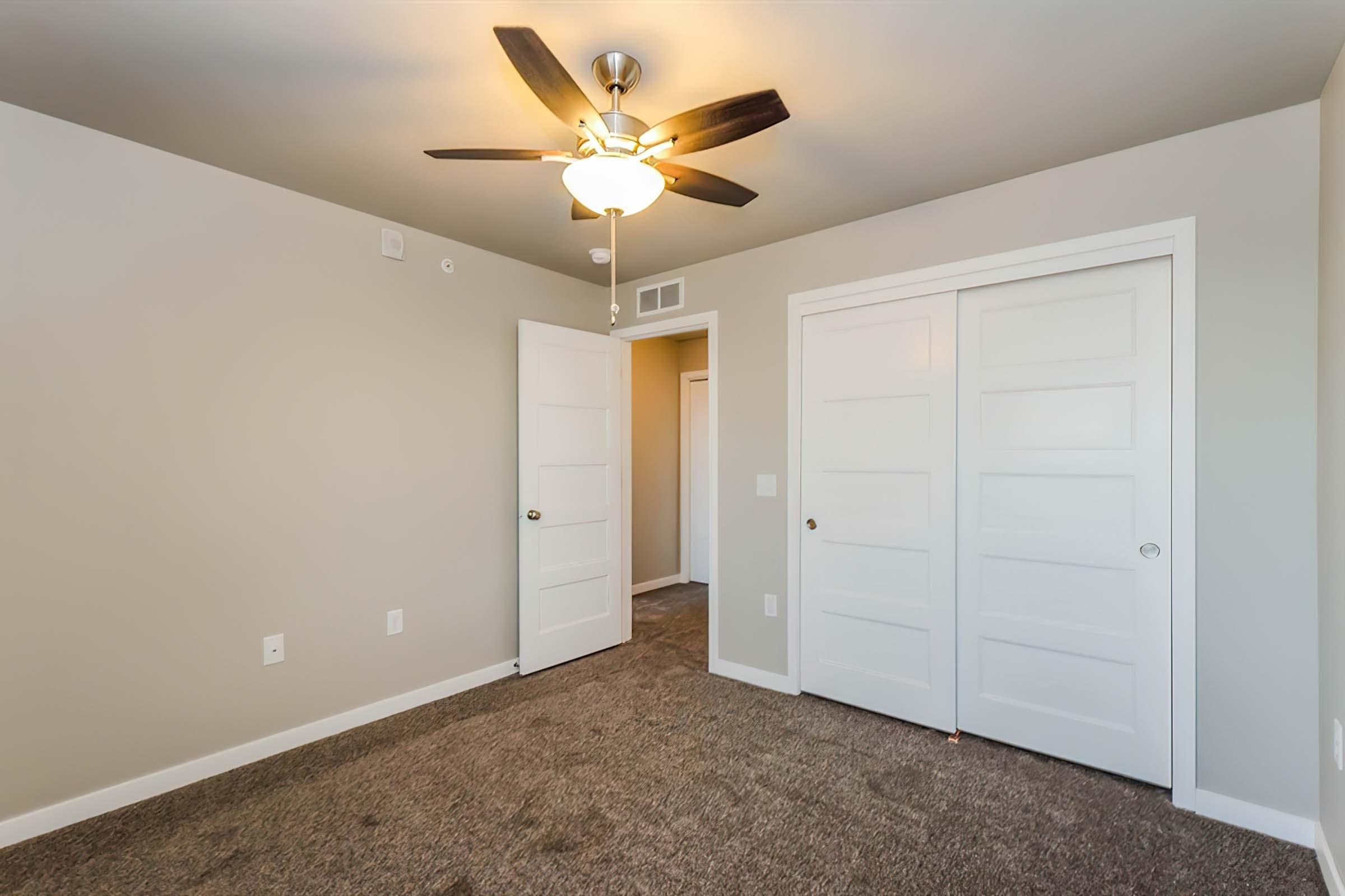 A well-lit bedroom featuring a ceiling fan, beige walls, and carpeted flooring. Two white double doors lead to a closet. The room has a neutral color scheme and a small hallway entrance visible in the background.