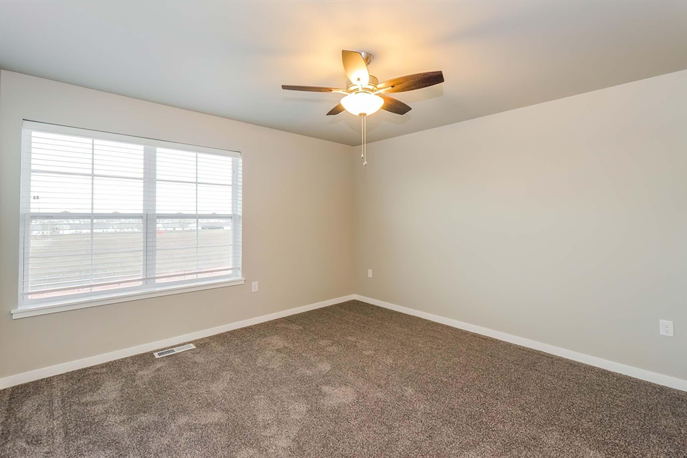 A clean, empty room featuring light gray walls and a ceiling fan. Natural light comes through a large window with white blinds, overlooking a view outside. The floor is covered in plush, beige carpet, creating a warm and inviting atmosphere.