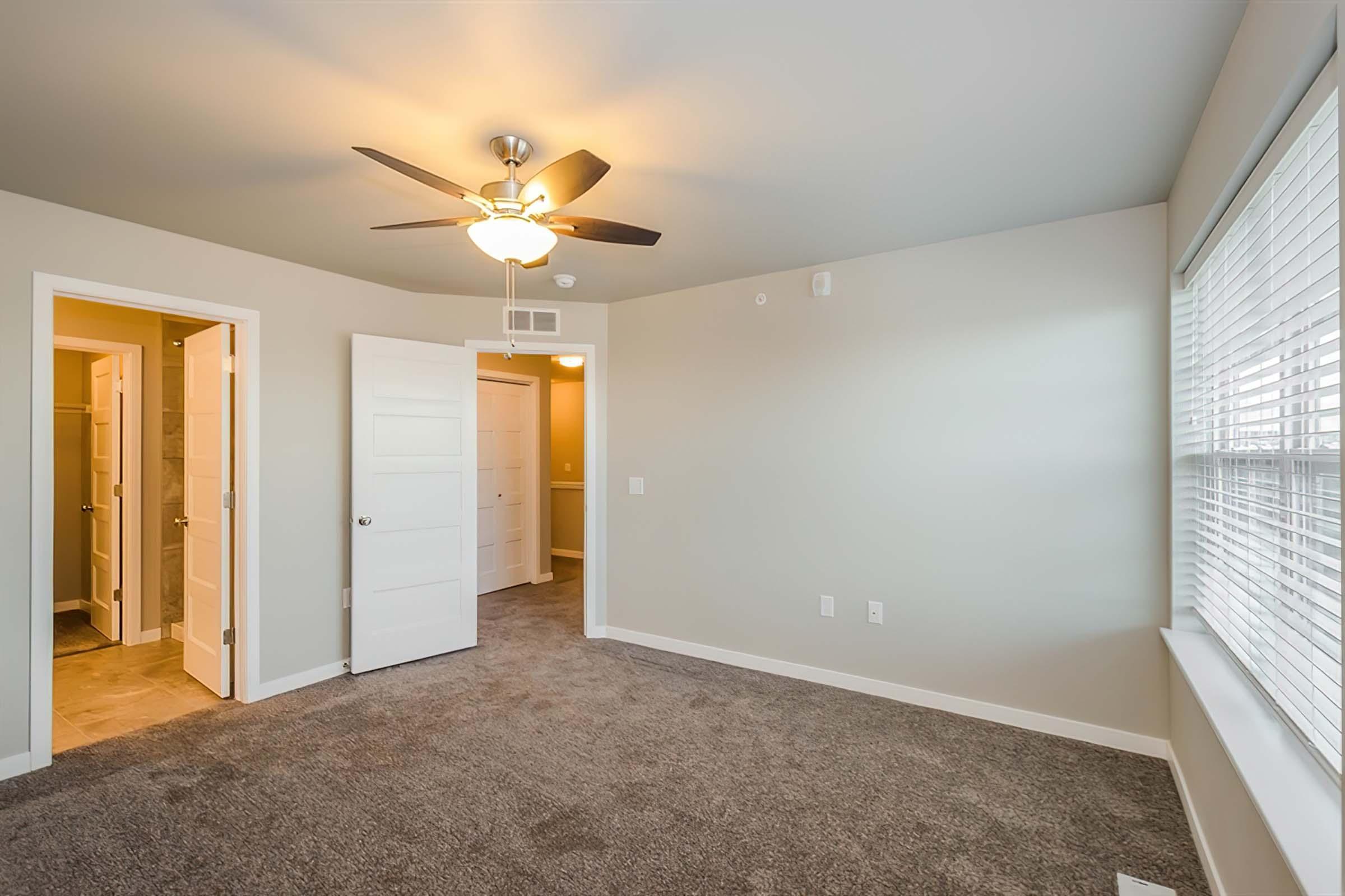 A well-lit room featuring a ceiling fan, carpeted flooring, and beige walls. The room has a large window and two doorways leading to adjacent spaces, suggesting a cozy, modern living area. The doors are closed, and the overall ambiance is neutral and inviting.