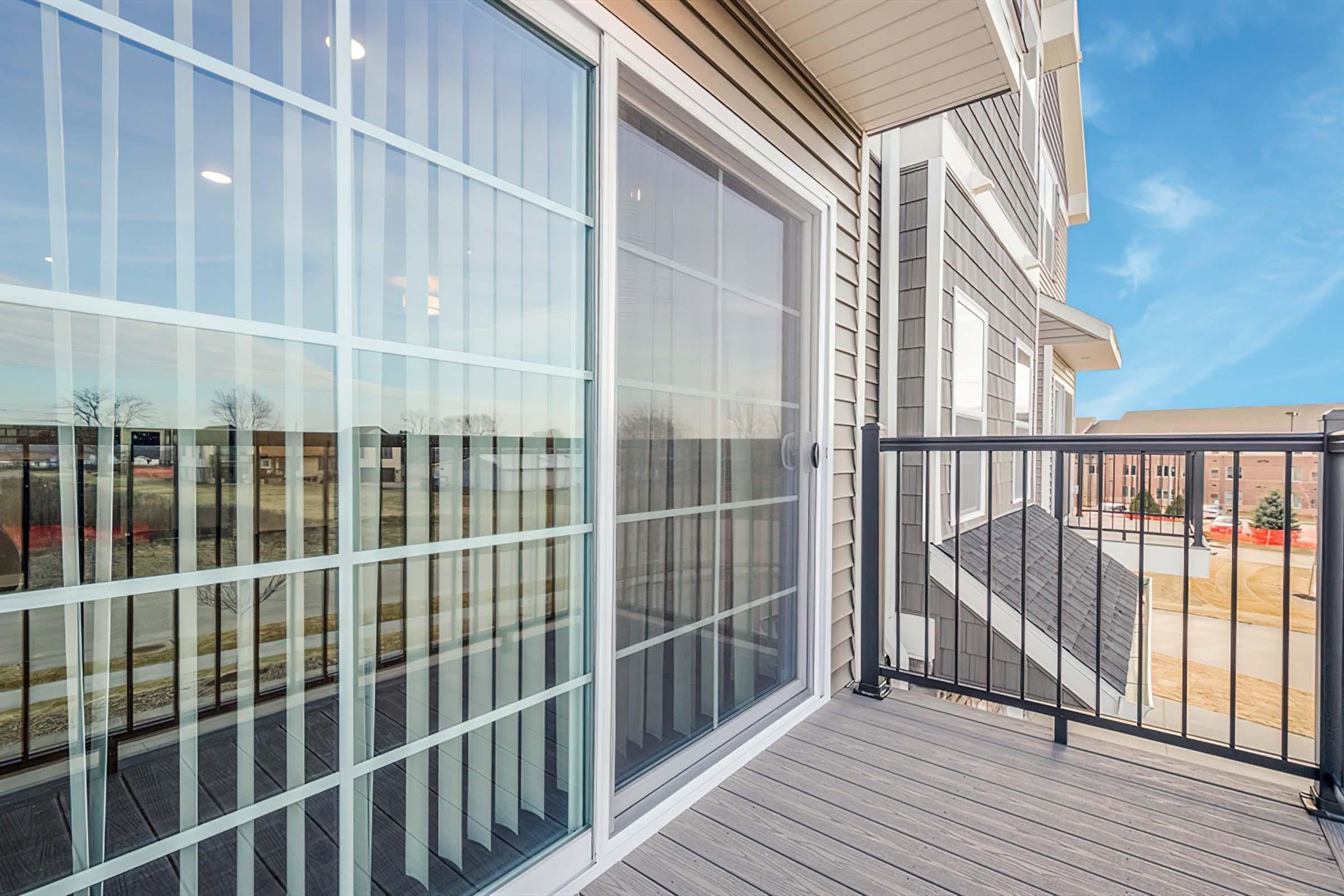 A balcony view with glass sliding doors featuring a grid pattern, overlooking a simple landscape. The deck is made of composite material, and the building has a modern exterior design. Clear skies are visible in the background, enhancing the bright ambiance of the scene.