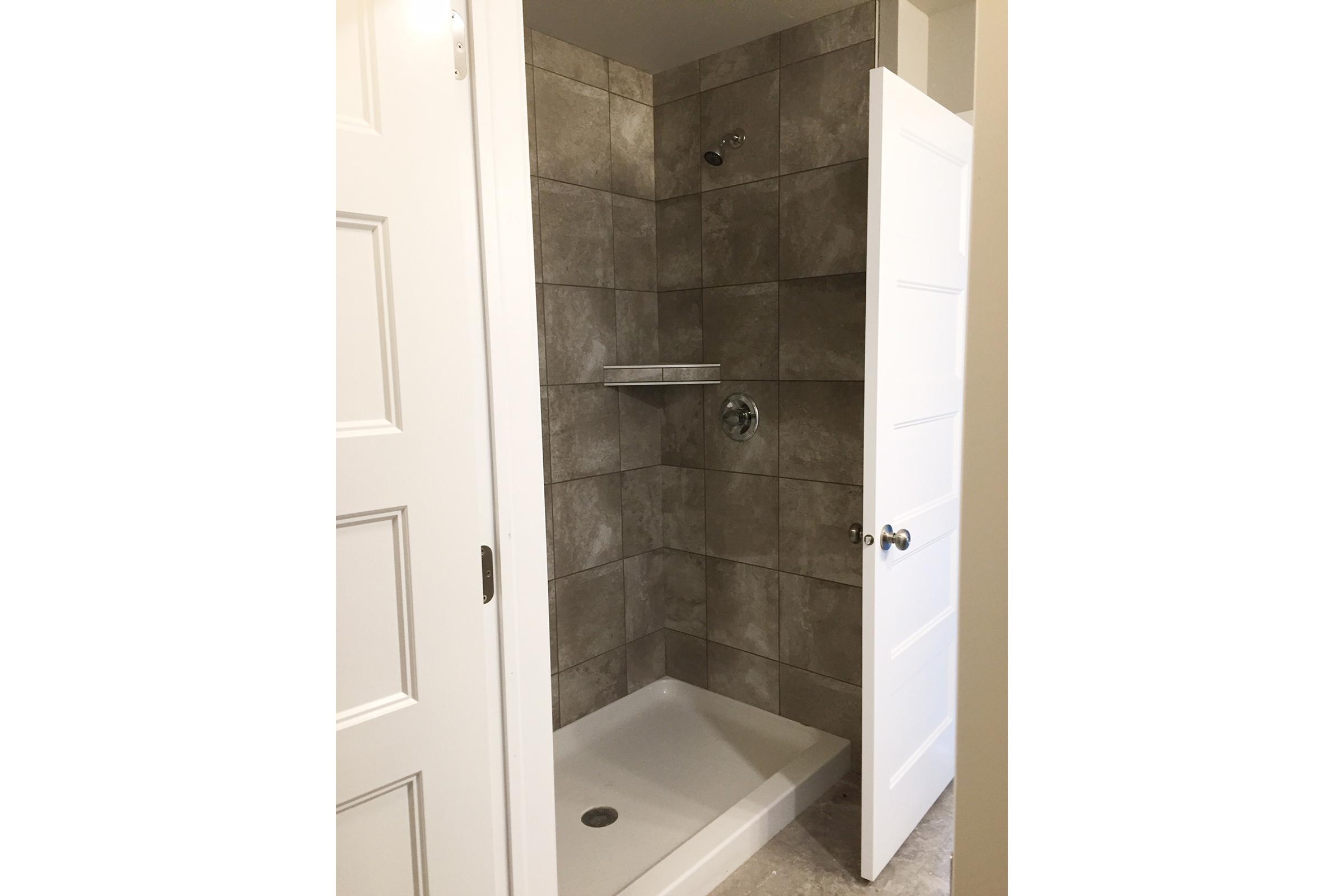 A modern, empty shower stall featuring gray tiled walls, a showerhead, and a small shelf. The shower is enclosed by a white door, which is slightly ajar, revealing a clean and minimalist design. The floor is finished with light-colored tiles, enhancing the contemporary look of the space.