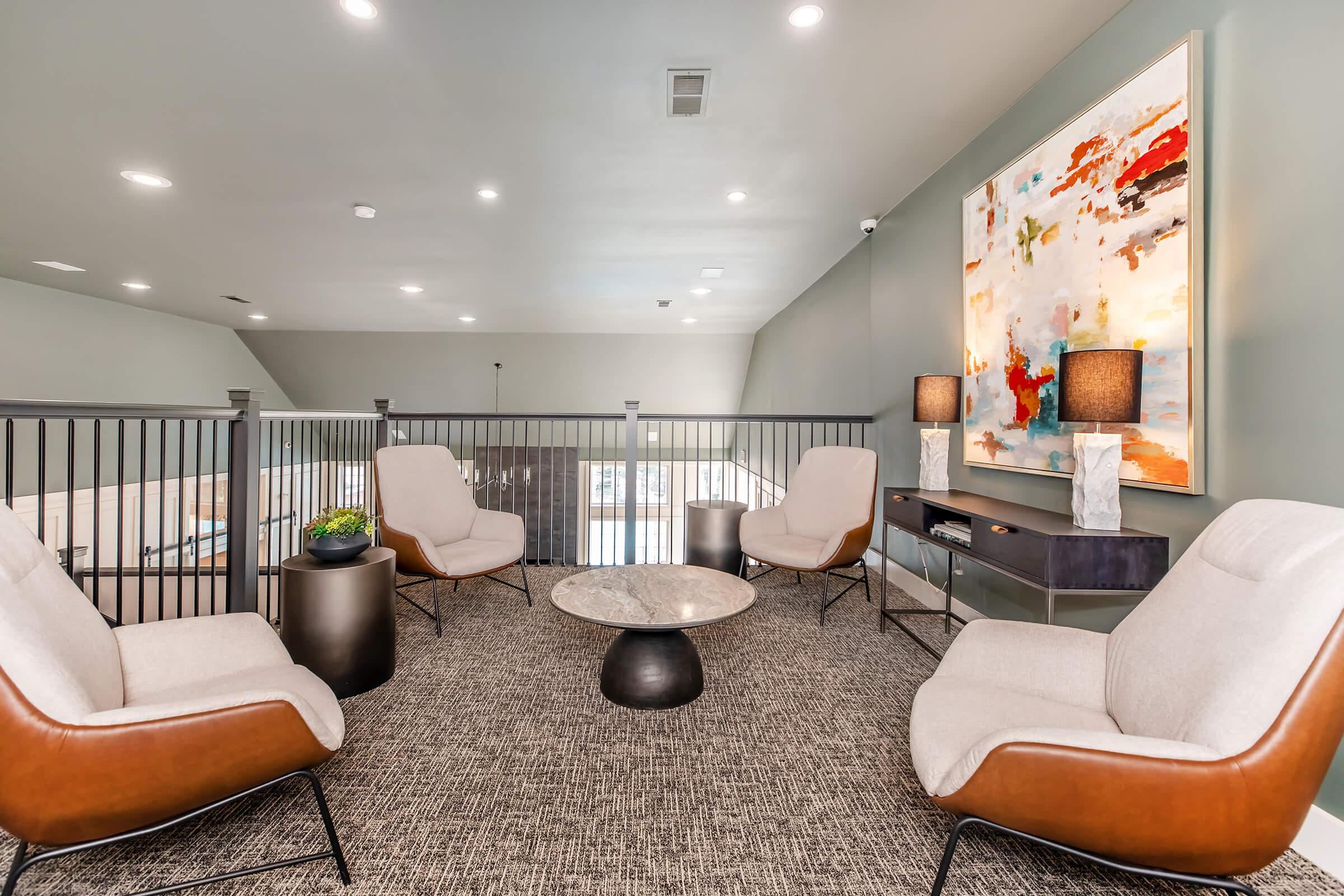 A modern lounge area featuring four comfortable armchairs, a round marble coffee table, and a stylish side table. The walls are adorned with abstract art, and soft lighting creates a welcoming ambiance. A railing leads to an upper level, enhancing the spacious feel of the room.