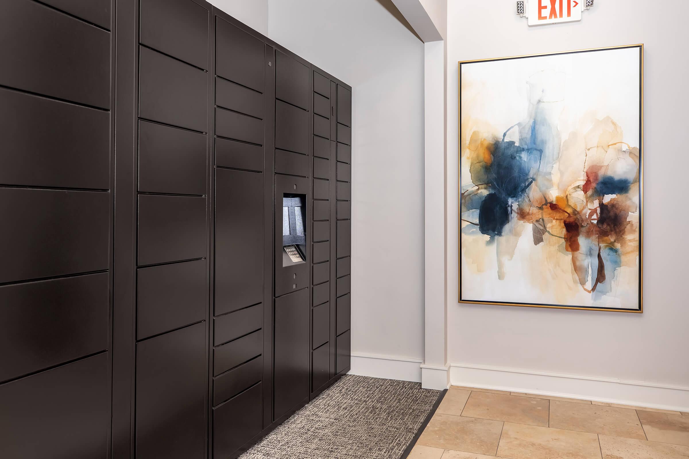 A modern hallway featuring a row of dark, sleek lockers on one side and a large abstract painting with blue, orange, and beige hues on the other. The floor is tiled, and there is a textured area rug in front of the lockers. An exit sign is visible above.