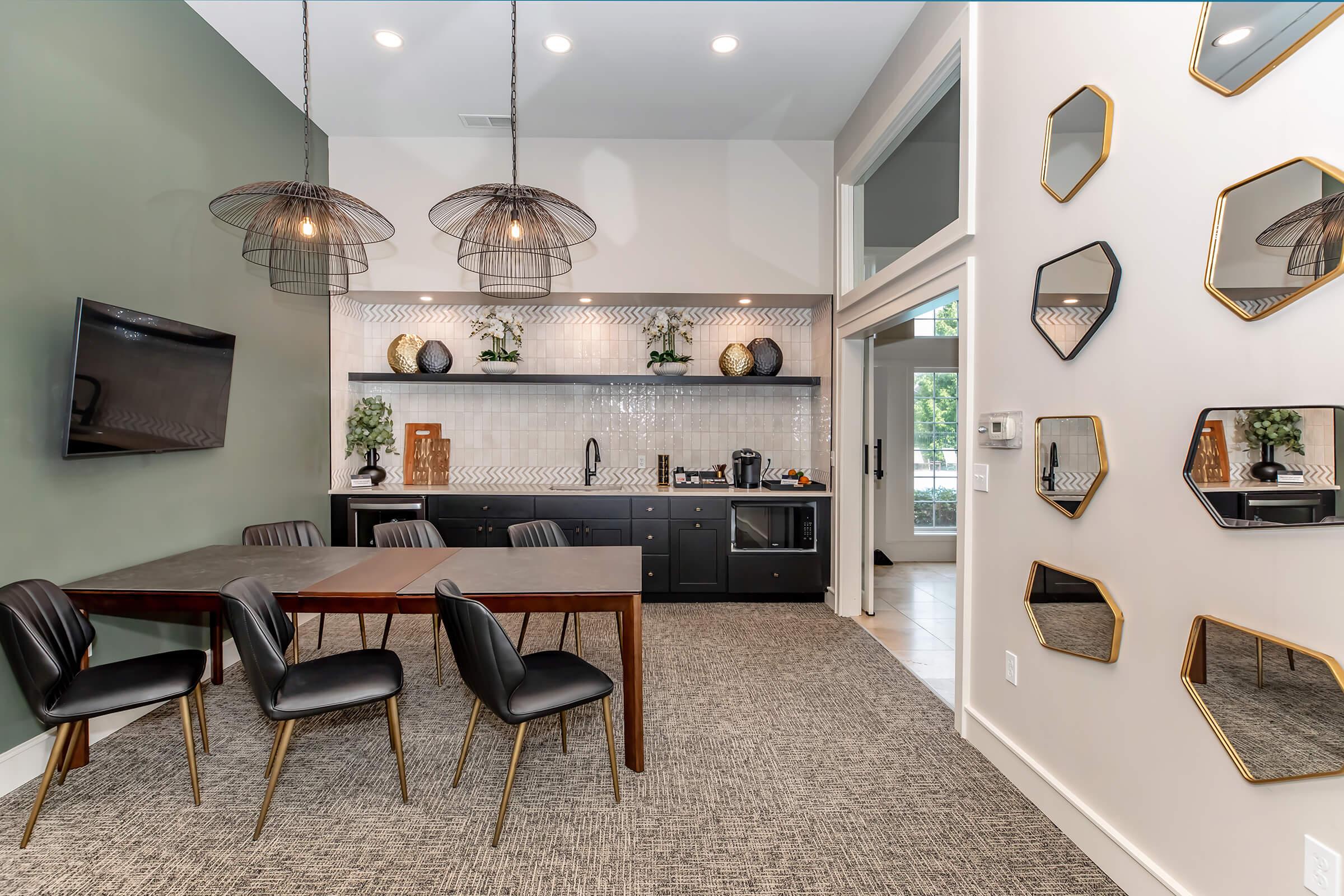 Modern kitchen and dining area featuring a wooden table with black chairs, decorative wall mirrors, and stylish pendant lighting. The kitchen has a sleek black cabinetry design with decorative plants and kitchen appliances, all set against a neutral color palette and textured wall.
