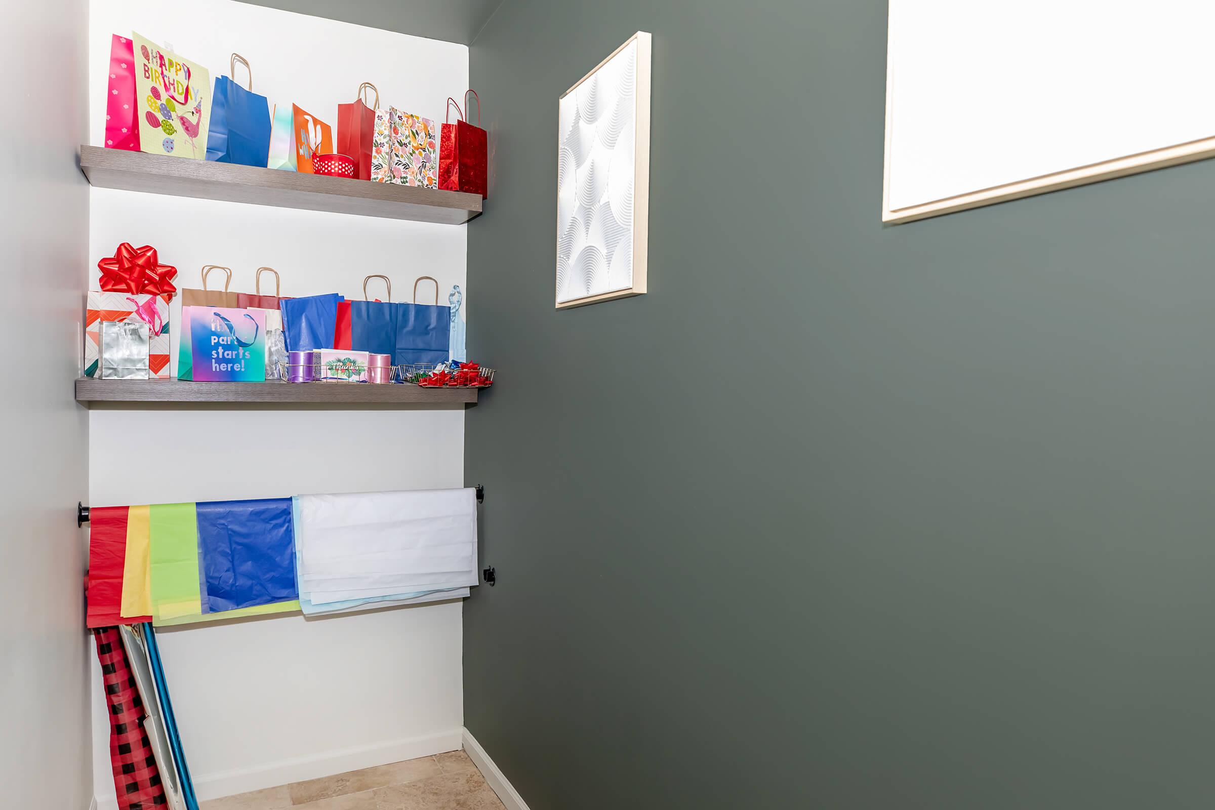 A small corner of a room featuring two shelves filled with colorful gift bags and wrapping supplies. The wall is painted a dark green, and there is a framed artwork on the wall. A variety of wrapping paper rolls can be seen hanging below the shelves, adding to the festive atmosphere.