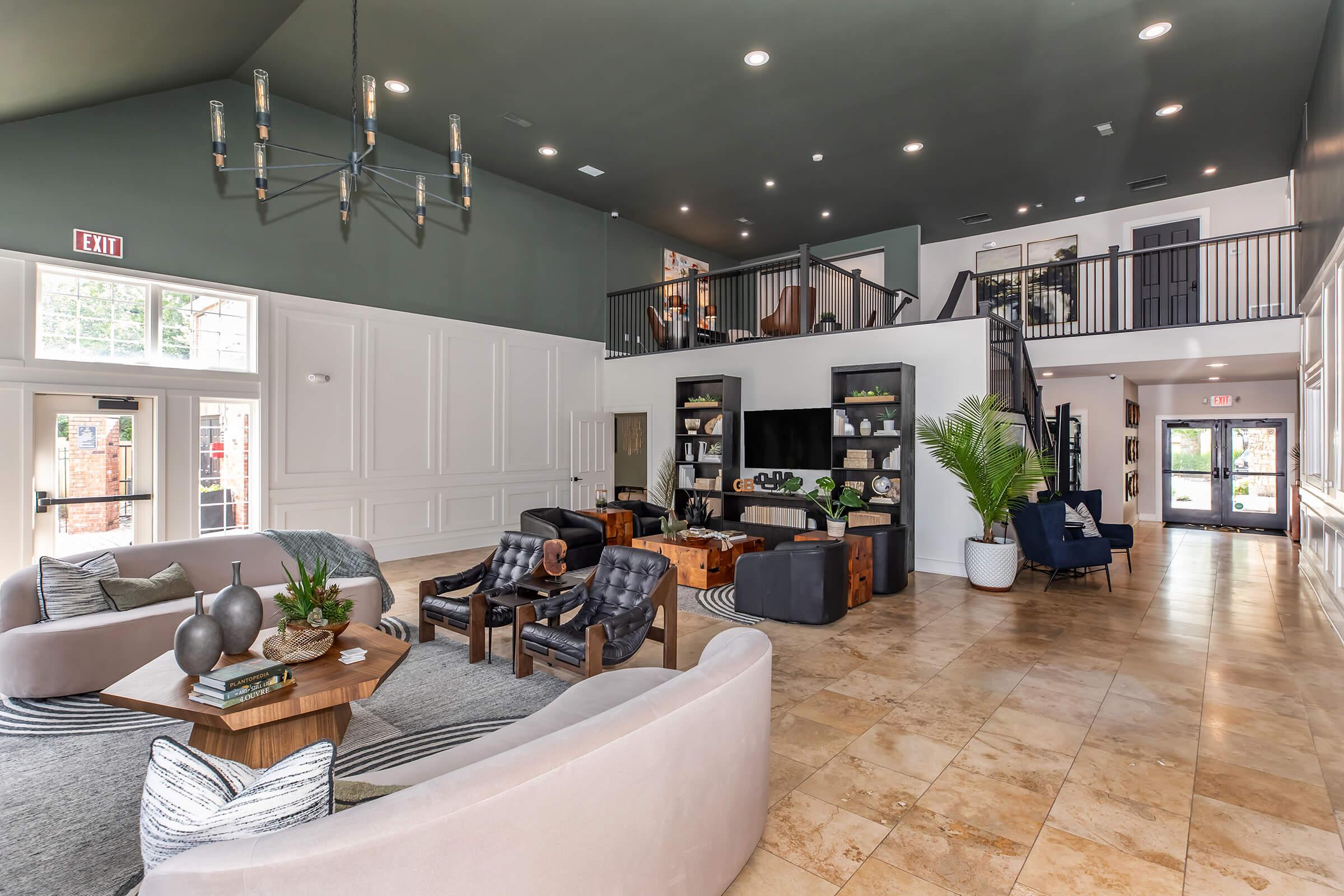 A spacious and modern lobby area featuring stylish furniture, including plush sofas and armchairs. The room has a high ceiling with elegant lighting, large windows that let in natural light, and decorative elements like plants and artwork. A staircase leads to an upper level with additional seating areas.