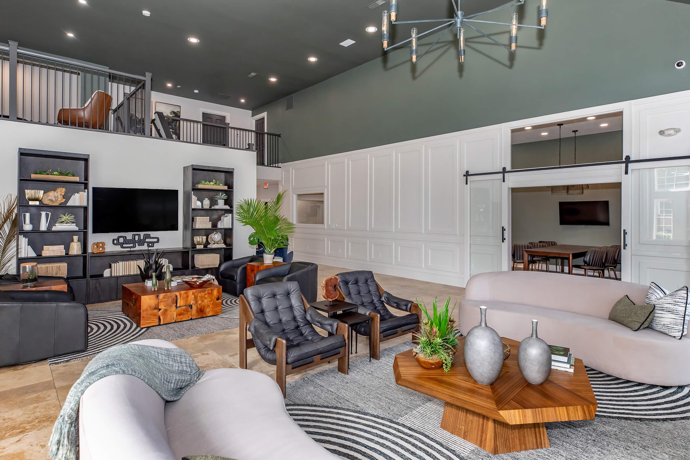 Modern lounge area featuring stylish furniture, including black armchairs and a light-colored curved sofa. The space has a large TV on a wall unit, decorative plants, and a wooden coffee table. Bright lighting and a high ceiling contribute to a spacious and inviting atmosphere.