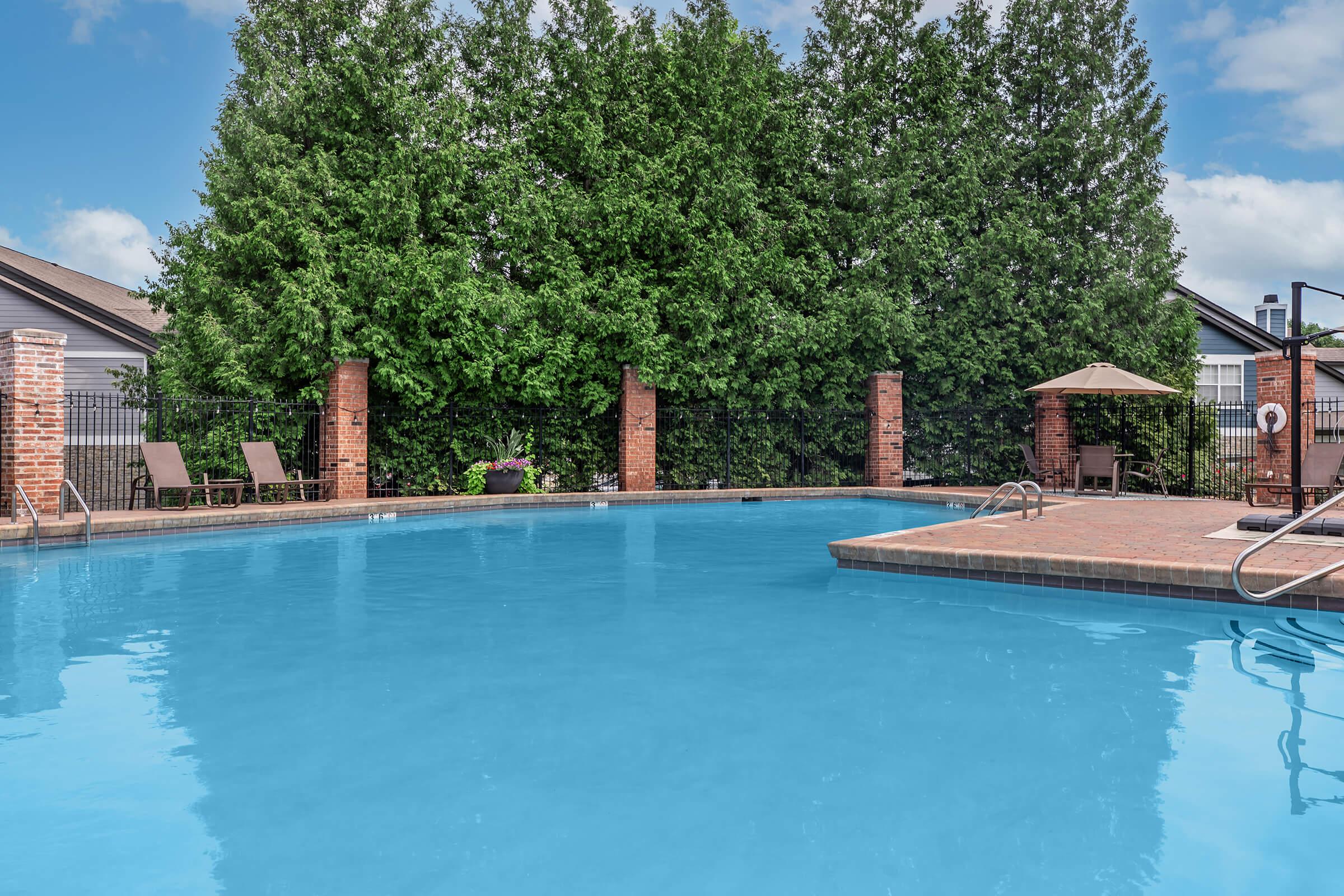 A tranquil outdoor swimming pool surrounded by greenery, featuring clear blue water. Brick columns and a fence provide a backdrop, with sun loungers positioned nearby. There's a shaded area with a canopy, and the scene is set against a bright blue sky. Ideal for relaxation and enjoyment.