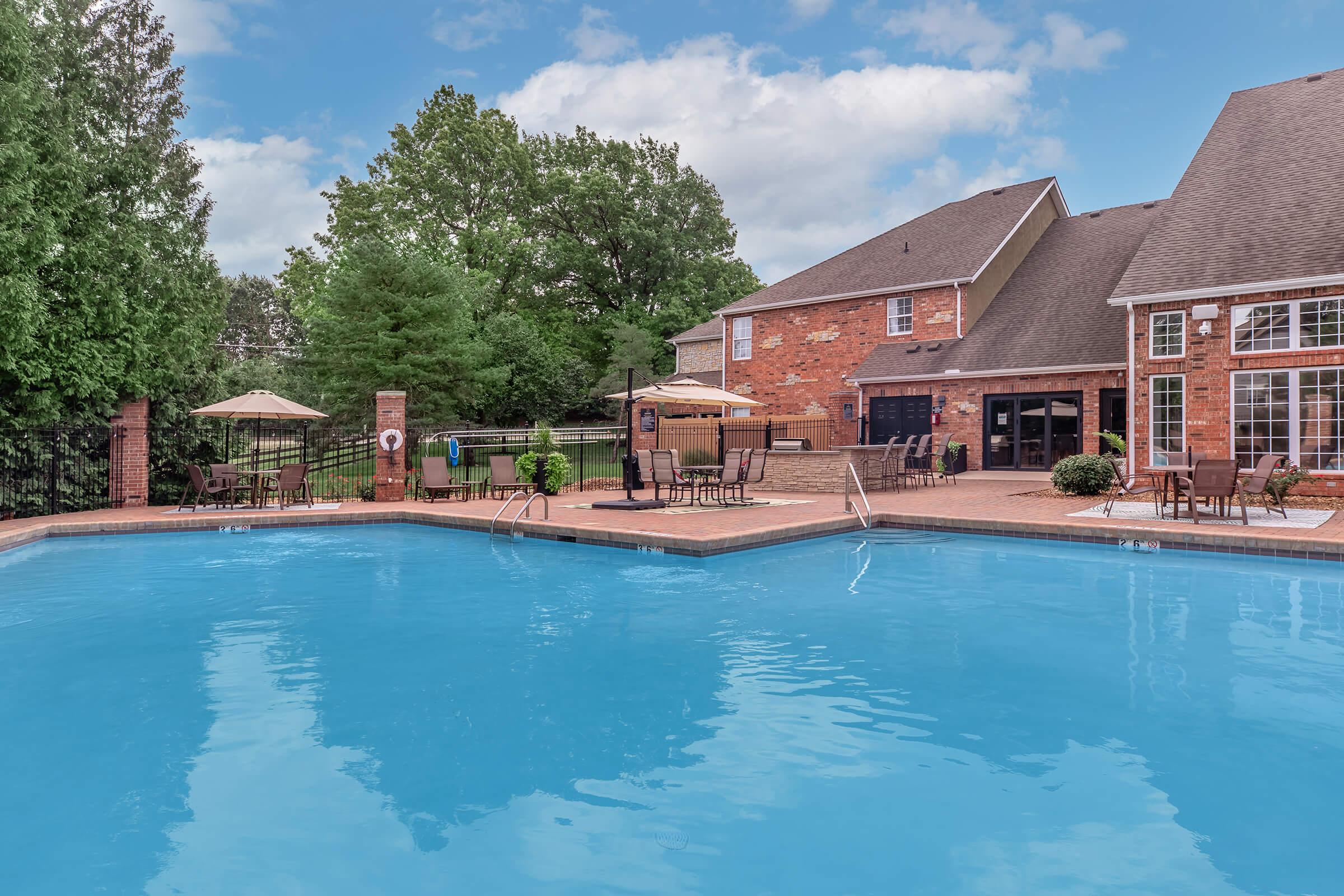 A bright outdoor swimming pool surrounded by a brick patio. There are lounge chairs and umbrellas near the pool, with green trees in the background. A brick building with large windows is adjacent to the pool area, creating a relaxing atmosphere for leisure and enjoyment.