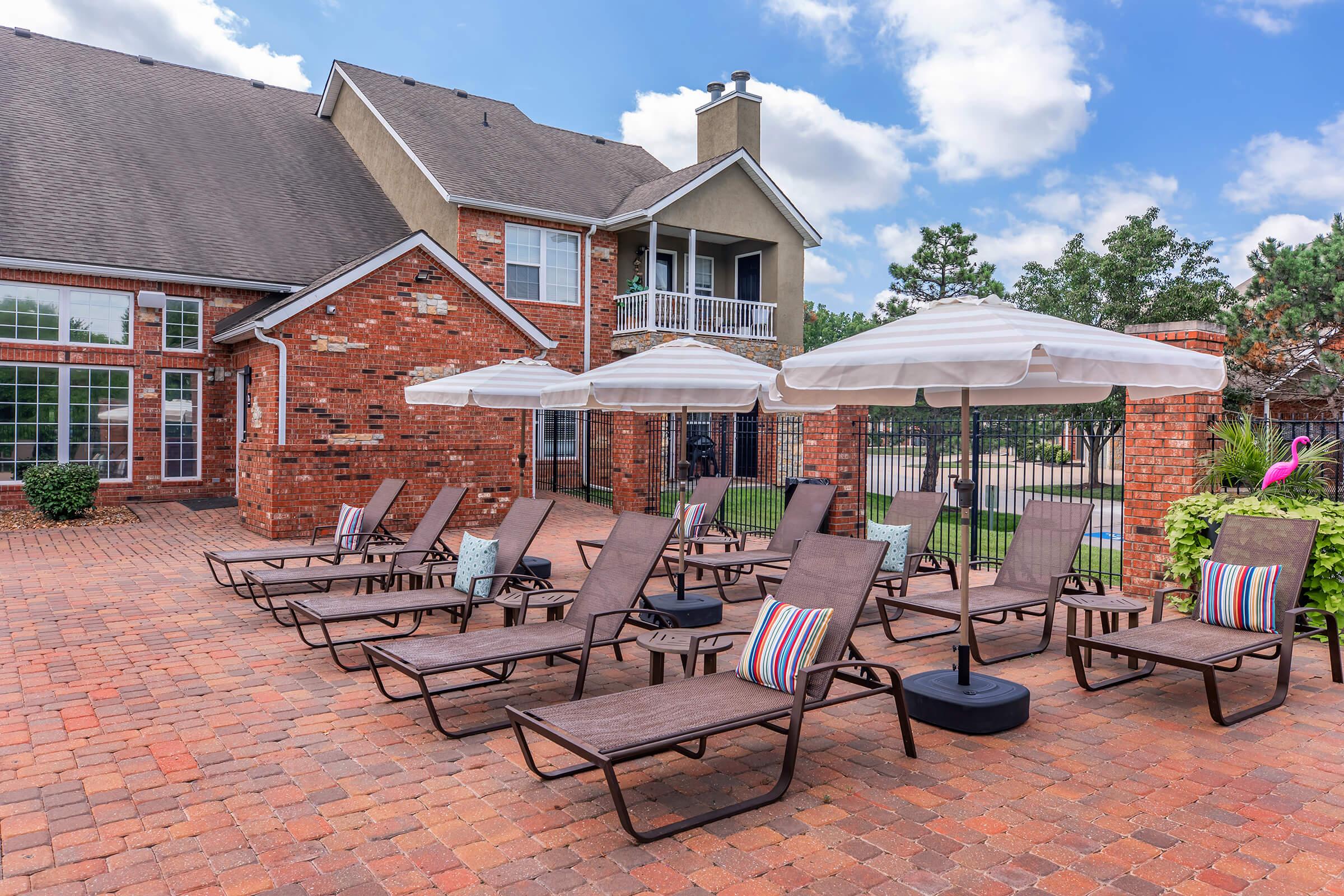 A spacious outdoor patio with several lounge chairs and colorful pillows, surrounded by a brick walkway. Two large umbrellas provide shade, while a brick building with large windows and a balcony is visible in the background. Green trees and a fence complete the inviting scene under a blue sky with fluffy clouds.
