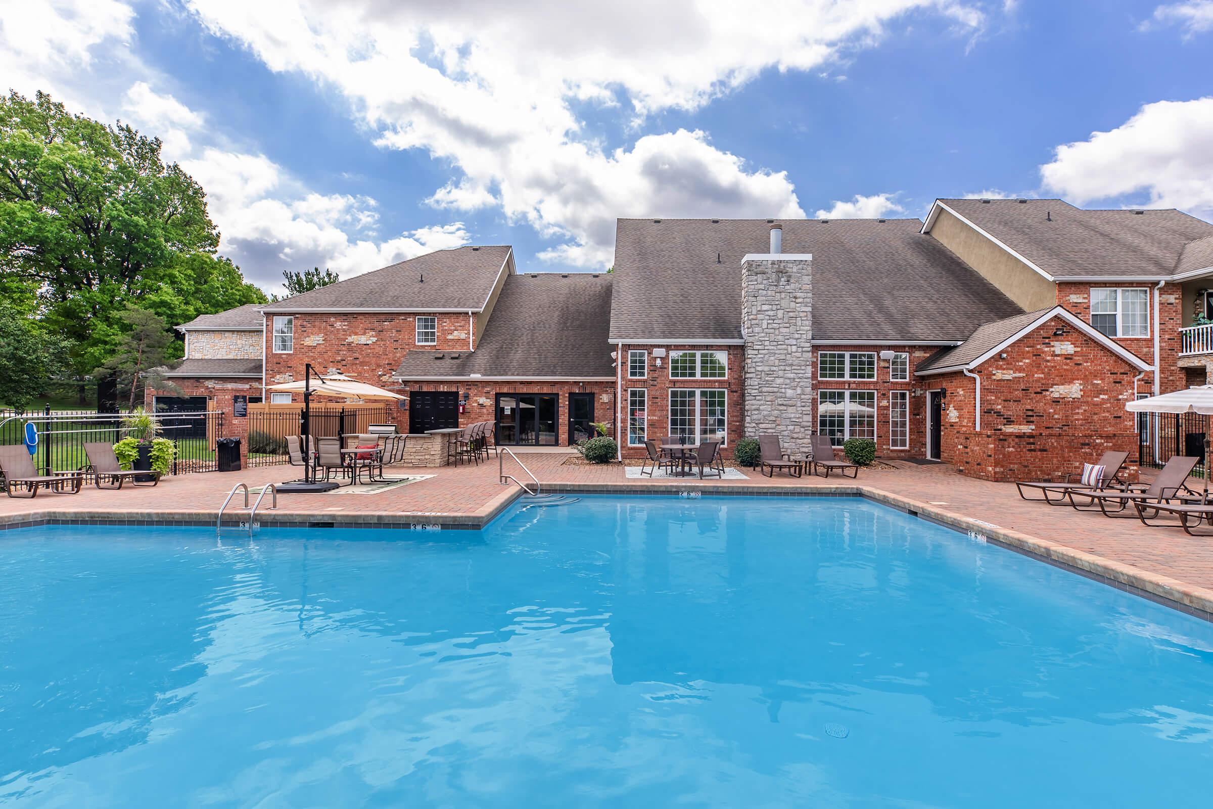 A swimming pool surrounded by lounge chairs, with a brick building in the background featuring large windows and outdoor seating areas. The sky is partly cloudy and the area is landscaped with trees.