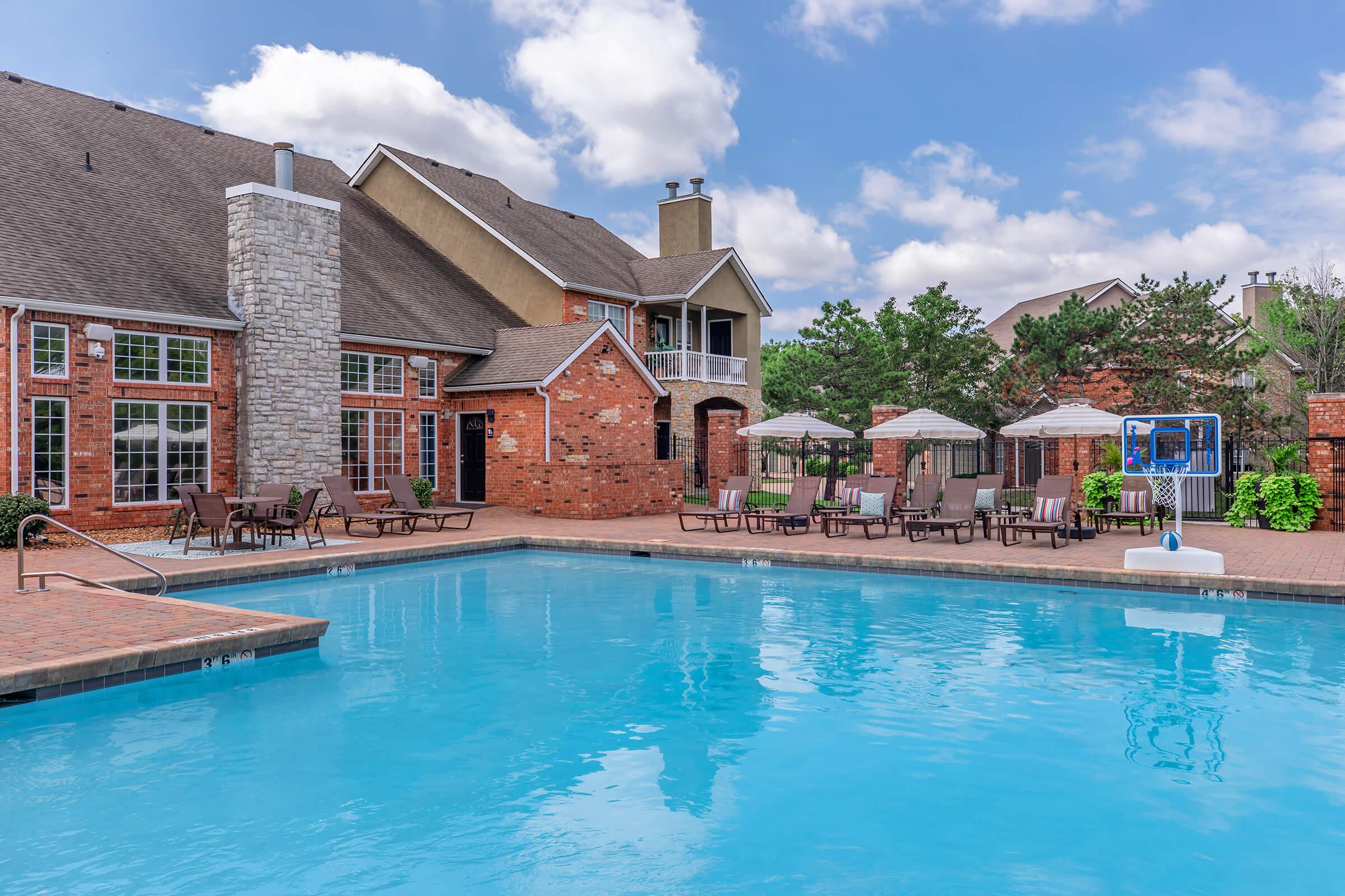 A well-maintained swimming pool surrounded by lounge chairs and umbrellas, with a brick patio. In the background, there's a multi-story residential building featuring stone and brick architecture, set against a partly cloudy sky with greenery surrounding the area.