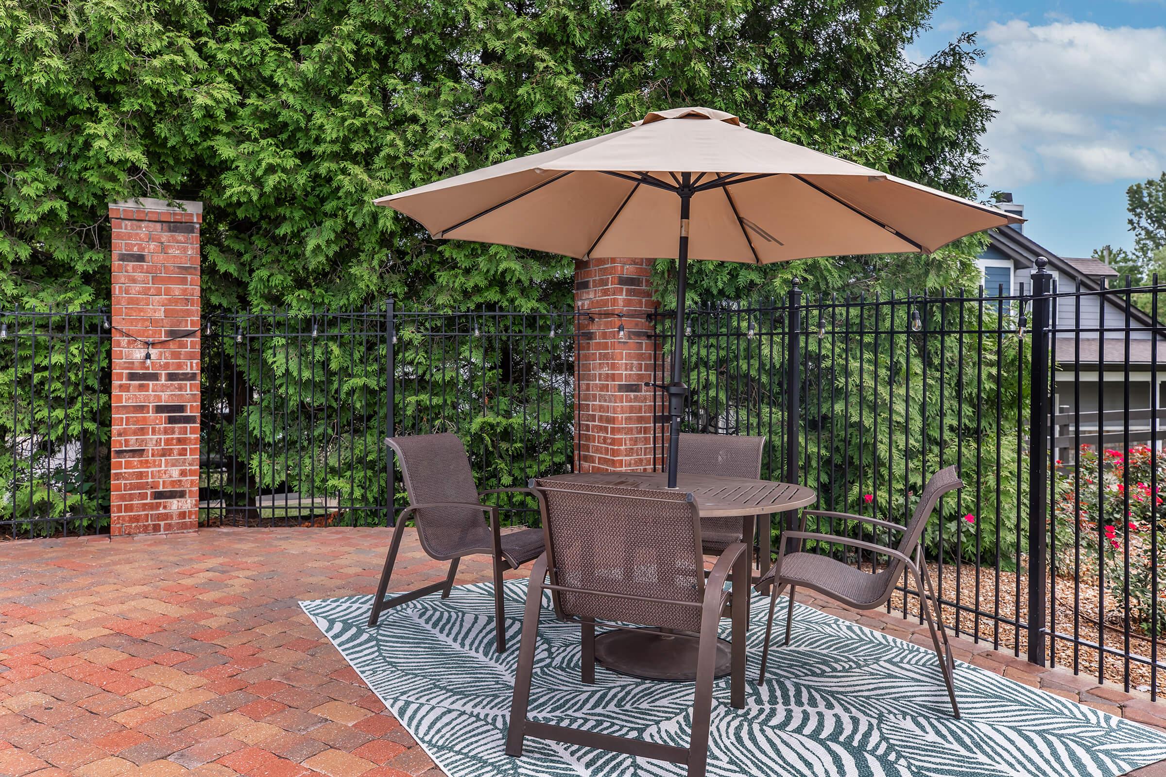 A cozy outdoor patio area featuring a round table with four chairs, shaded by a large tan umbrella. The floor is covered with a green and white patterned rug, and the background has tall greenery and a black fence, creating a tranquil setting.