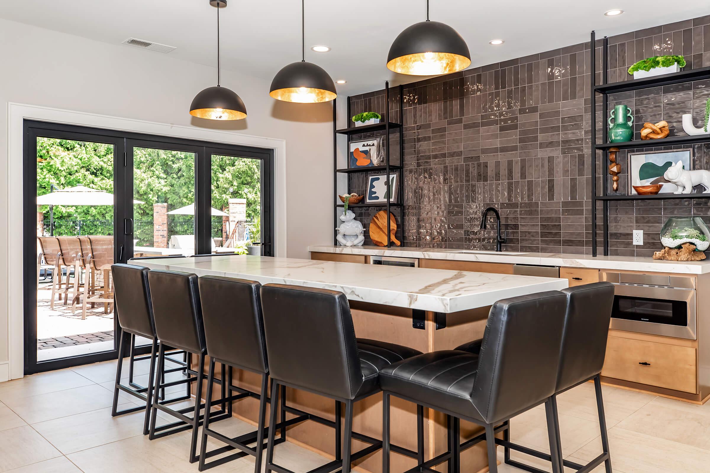 A modern kitchen featuring a large island with black bar stools, pendant lighting, and sleek cabinetry. The backsplash consists of dark tiles, and there are decorative elements on shelves. A view of a patio with outdoor seating is visible through glass doors.