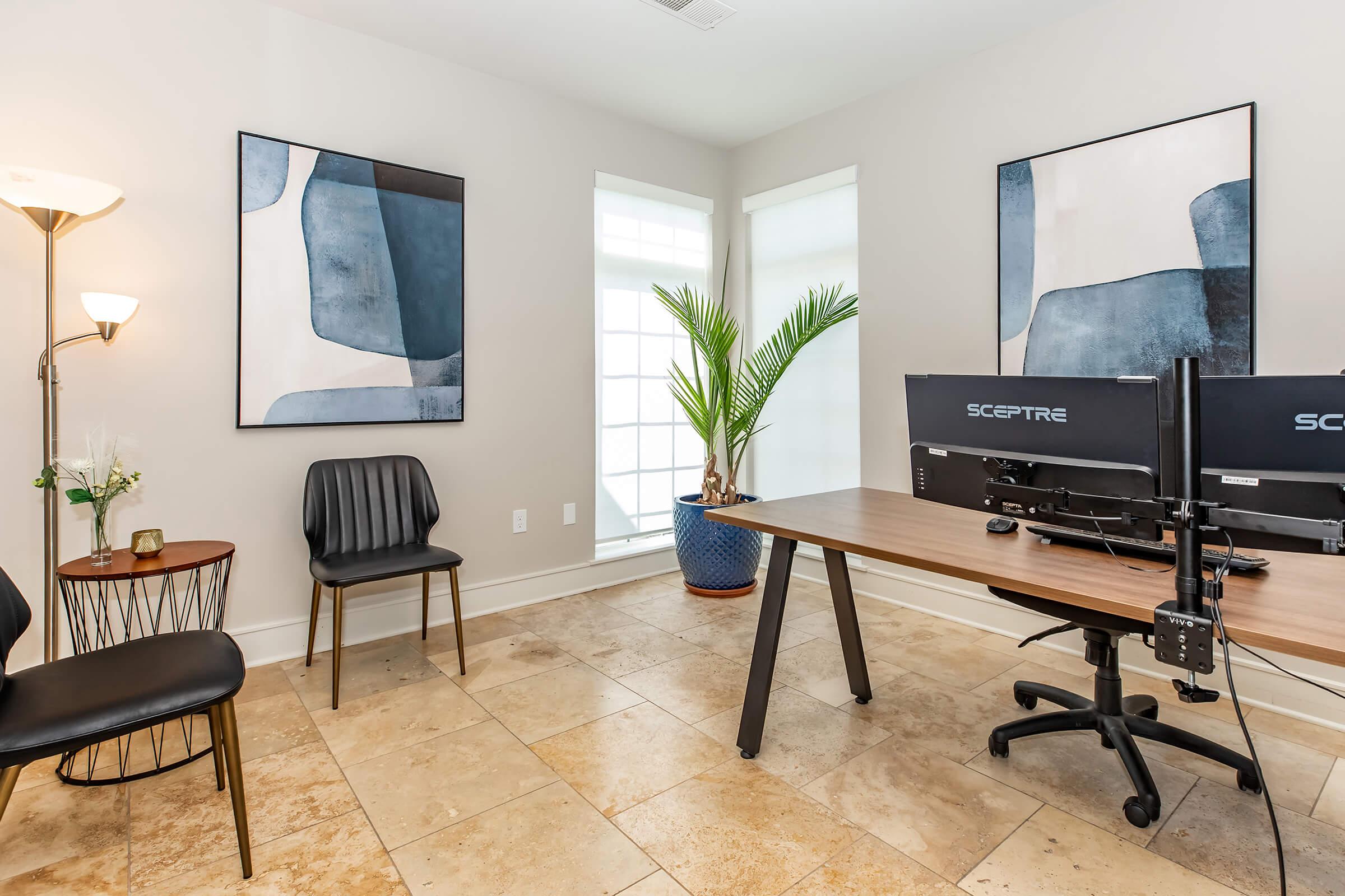 A modern office space featuring a wooden desk with dual monitors, a black chair, and two additional chairs. The room has light-colored walls, large windows allowing natural light, and two abstract art pieces hanging on the walls. A tall potted plant adds a touch of greenery to the space.