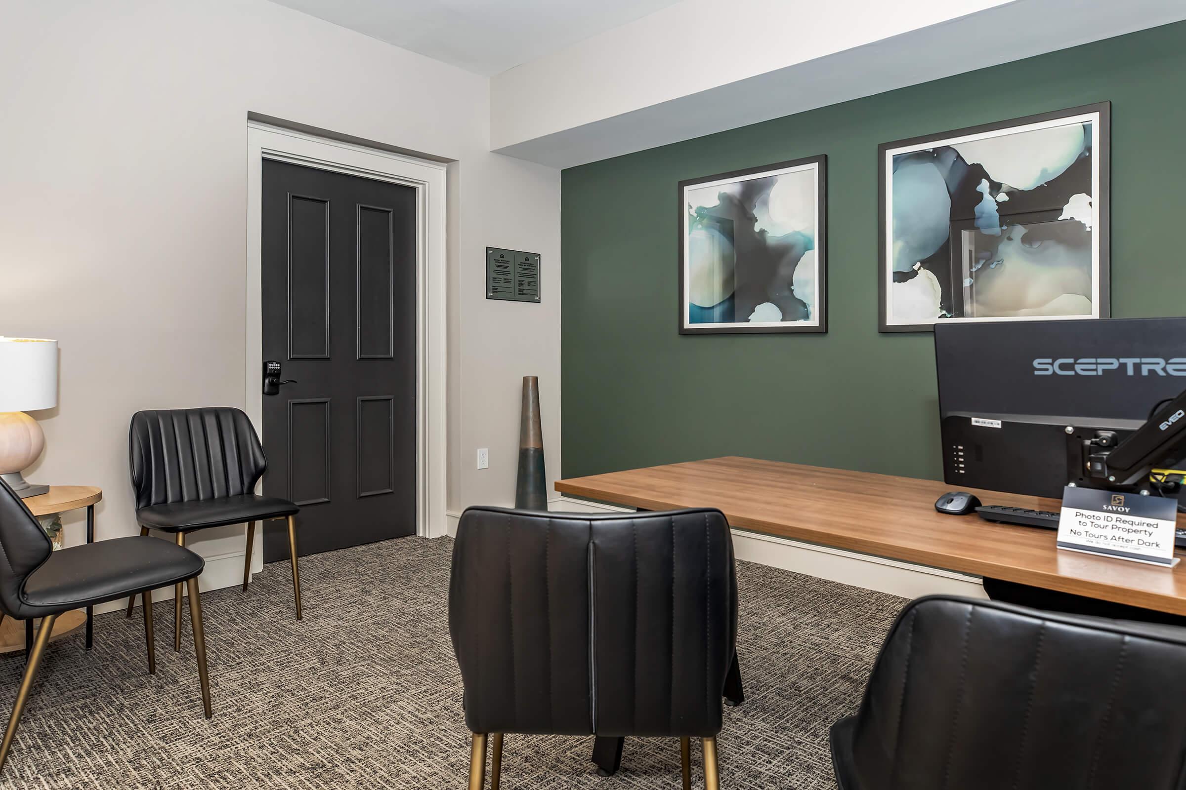 A modern office space featuring a wooden desk with a computer monitor, surrounded by stylish black chairs. The walls are painted in a muted green, and there are two abstract framed artworks. A door leads to another space, and there’s a decorative floor piece in the corner. Soft lighting creates a welcoming atmosphere.