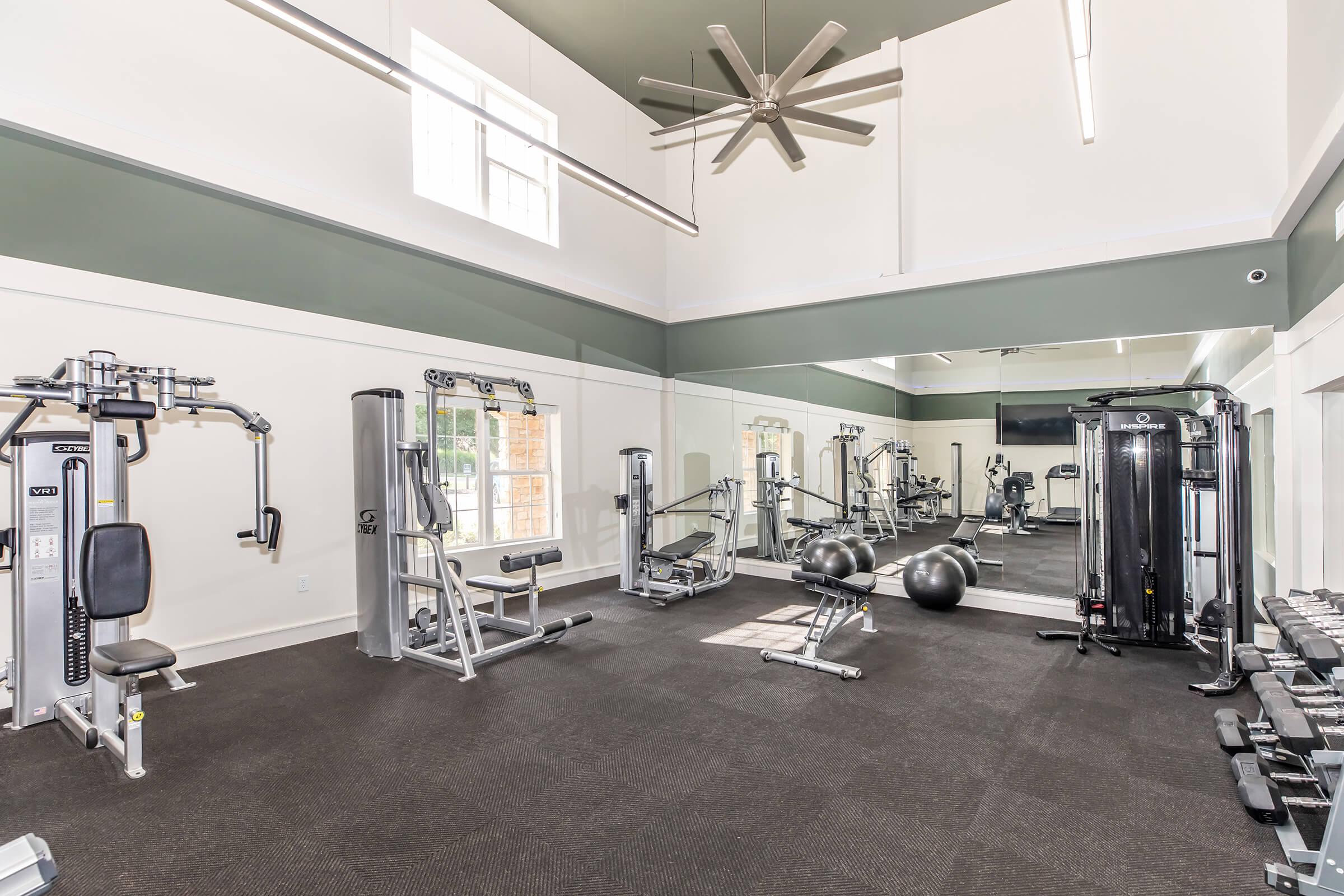 A modern gym interior featuring various exercise equipment including weight machines, free weights, and a large mirror. The space is well-lit with natural light coming through windows, and a ceiling fan is visible. The flooring is dark with a clean and organized layout.