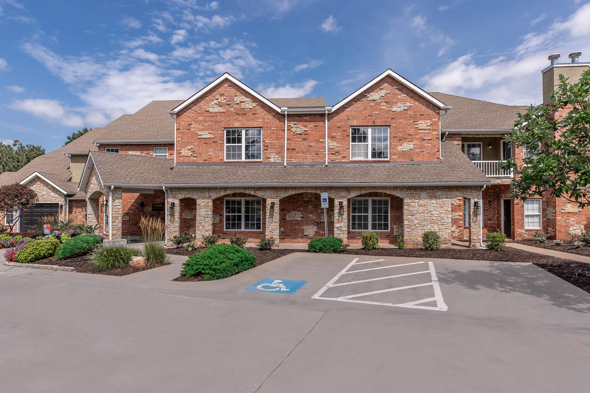 A two-story brick building with a porch, featuring a landscaped entrance with shrubs and flowers. A designated handicapped parking space is visible in the foreground. The sky is partly cloudy, creating a bright atmosphere around the property.