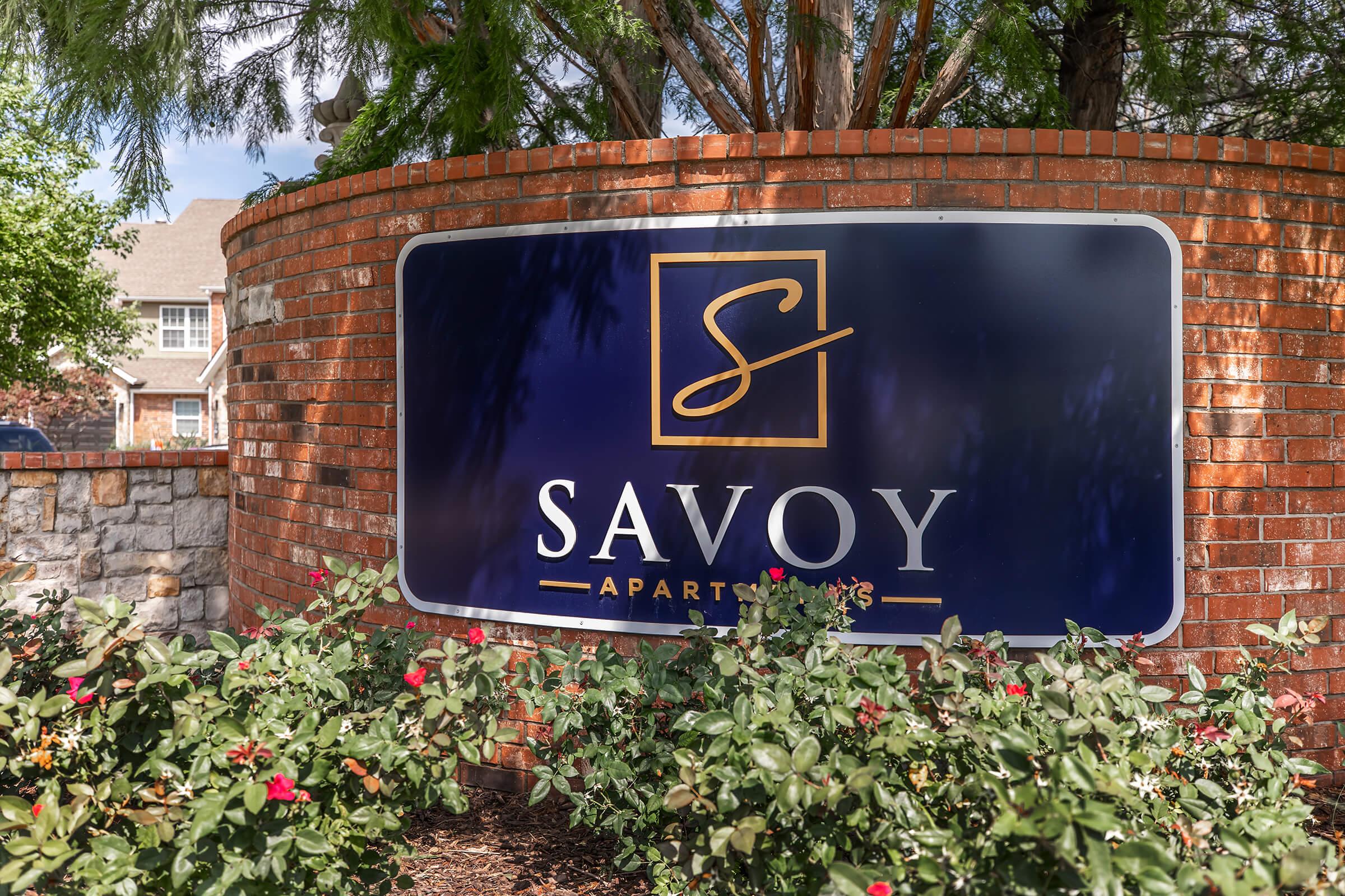Sign for Savoy Apartments featuring a stylized "S" logo. The sign is surrounded by brick and greenery, with blooming flowers below.