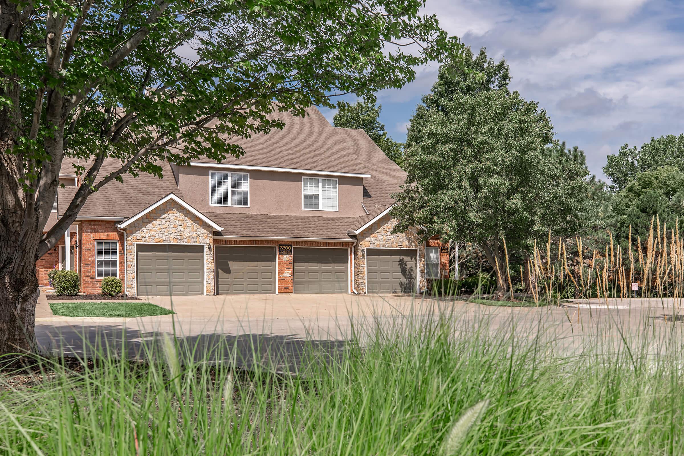 A spacious two-story home with a peaked roof and multiple garage doors, surrounded by green grass and trees. The driveway is clear, and the home features brick and neutral siding. In the background, there are tall grasses and shrubs, with a partly cloudy sky above.