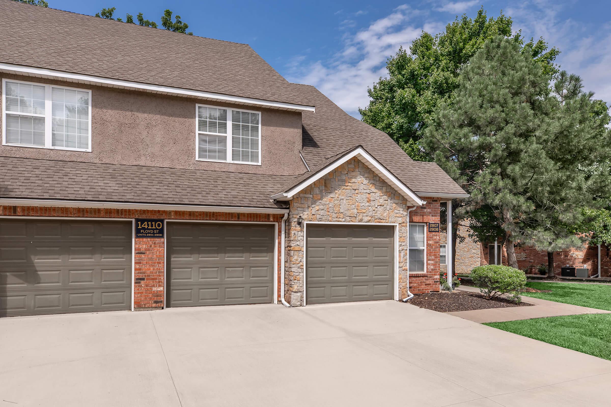 Two garage doors of a residential building with a stone and brick exterior. The building features a sloped roof and several windows. Lush green trees and a well-maintained lawn can be seen in the background under a clear blue sky.