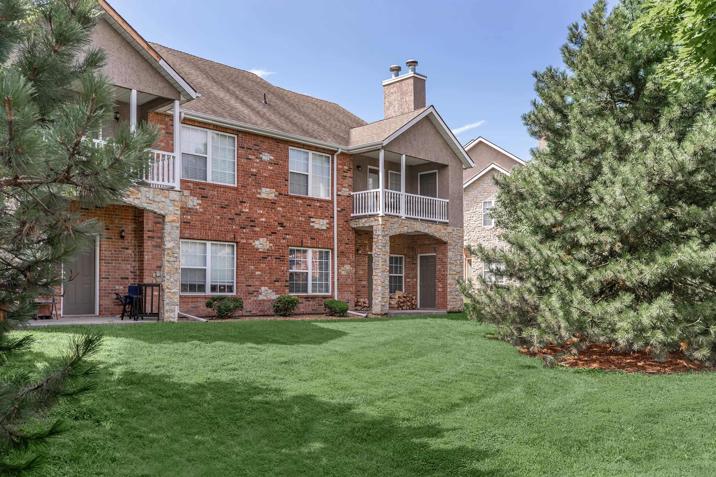 A view of a multi-story brick apartment building surrounded by lush green grass and trees. The building features multiple windows and balconies, with a neatly landscaped yard. Clear blue sky overhead adds to the pleasant atmosphere.