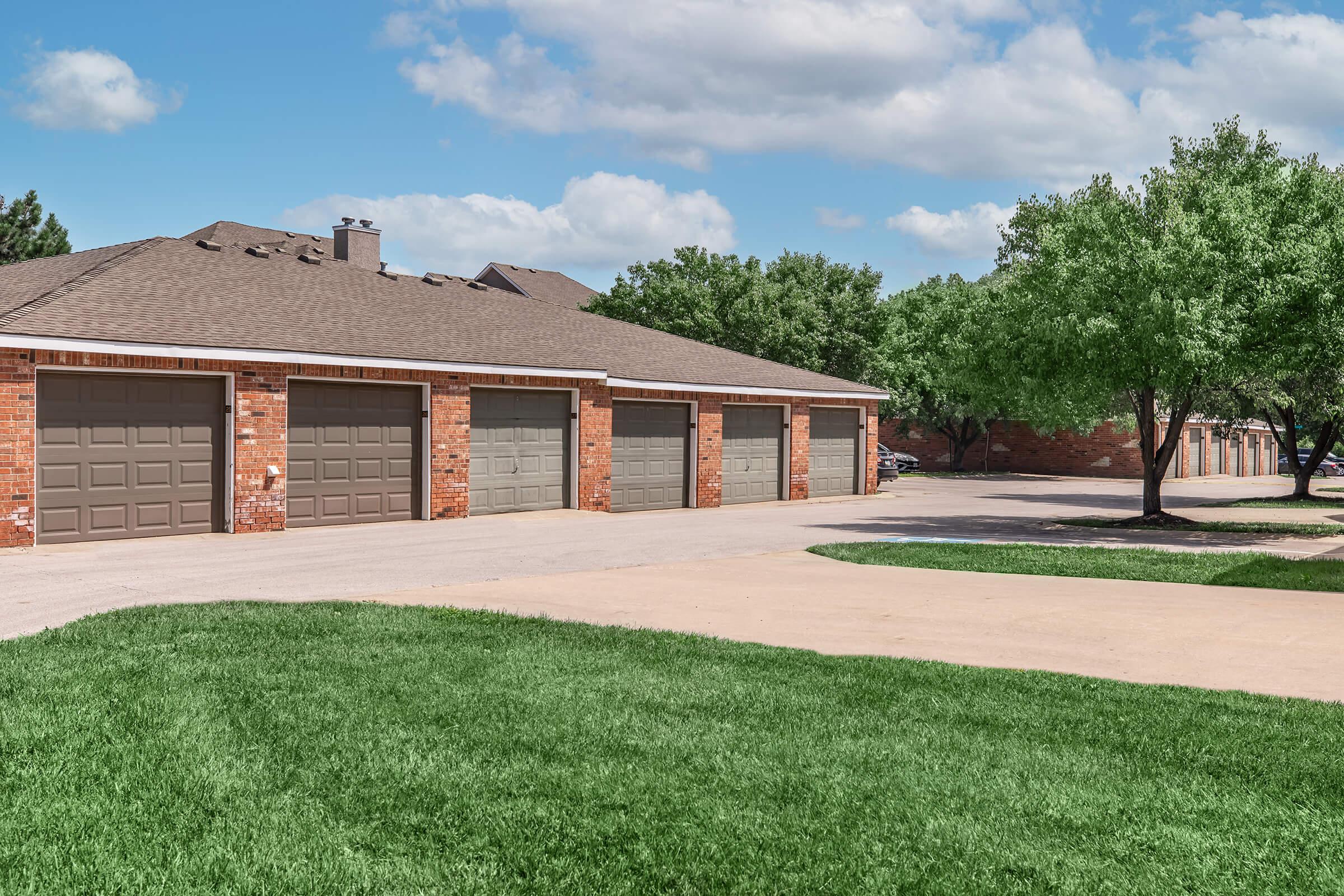 A residential building featuring multiple garages with brown doors, set against a backdrop of green grass and trees. The sky is partly cloudy, creating a bright and inviting atmosphere. The building has a brown shingle roof and is located in a suburban area.