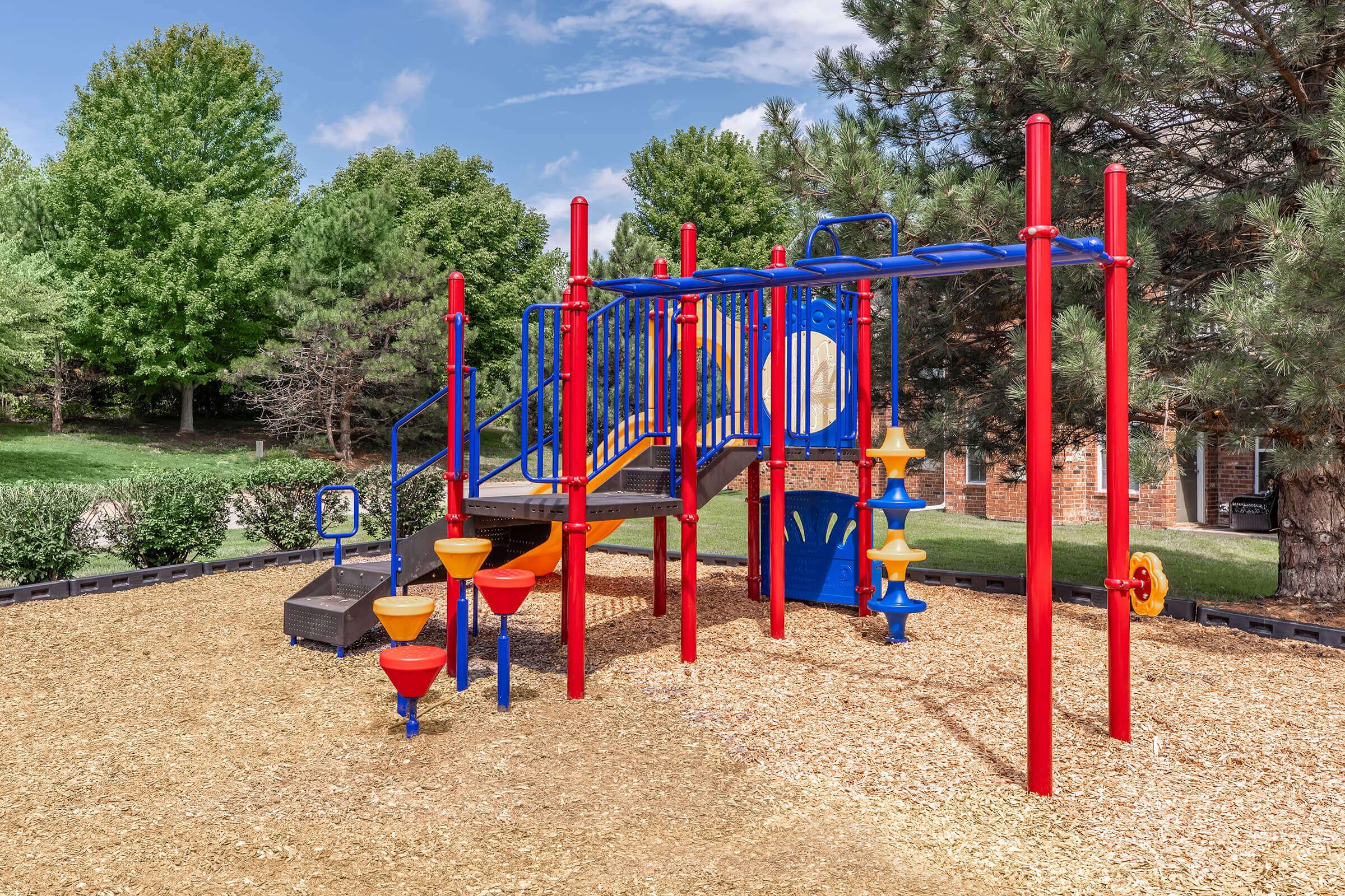 Colorful playground equipment featuring slides, climbing structures, and interactive panels, surrounded by a grassy area and trees. The ground is covered with wood chips, providing a safe play surface. Ideal for children's recreational activities.