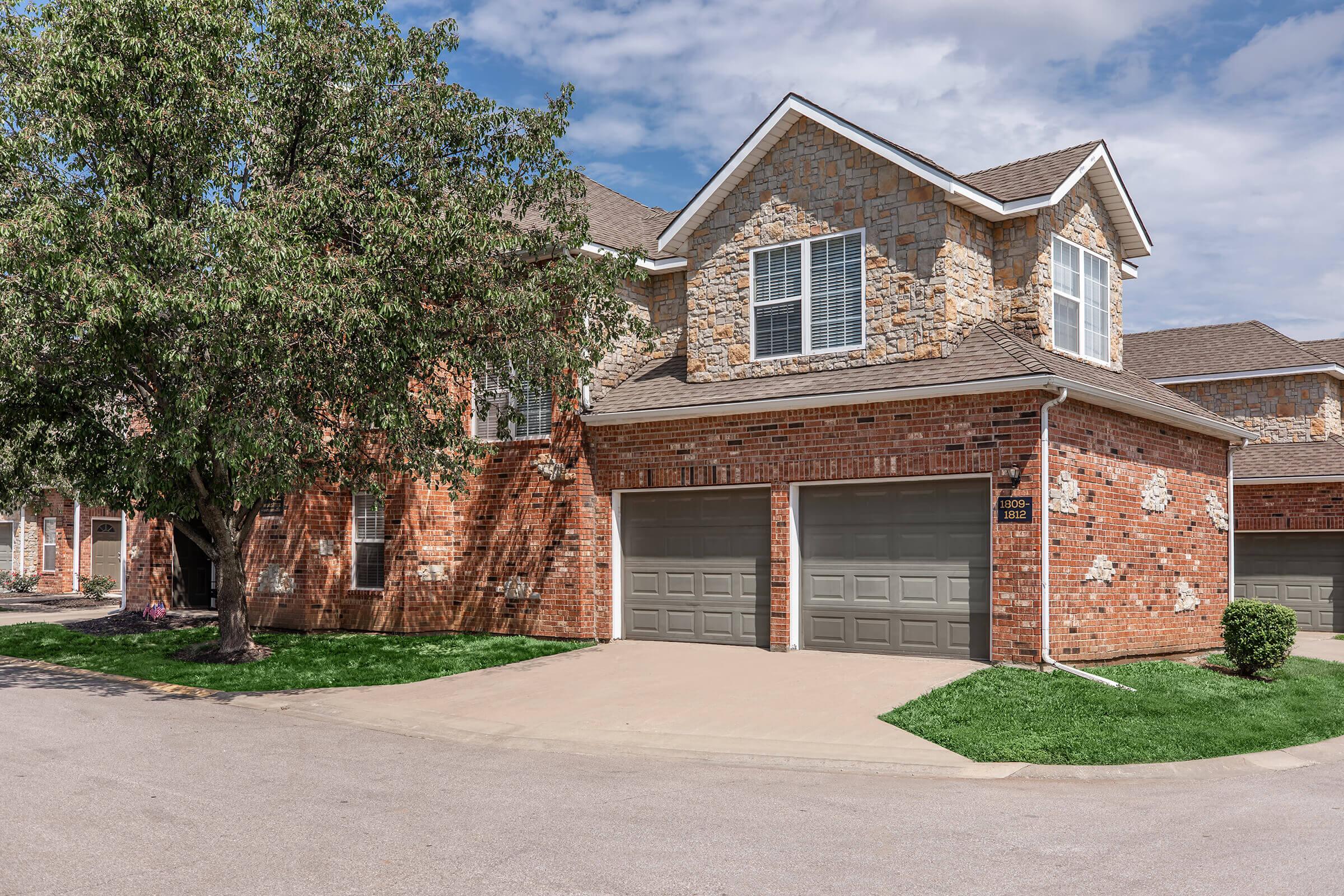 A residential building with a brick and stone exterior, featuring two garage doors on the front. The structure is set in a suburban area with a well-maintained lawn and a tree nearby. Part of a larger complex, the sky is partly cloudy, and the driveway curves around the corner.