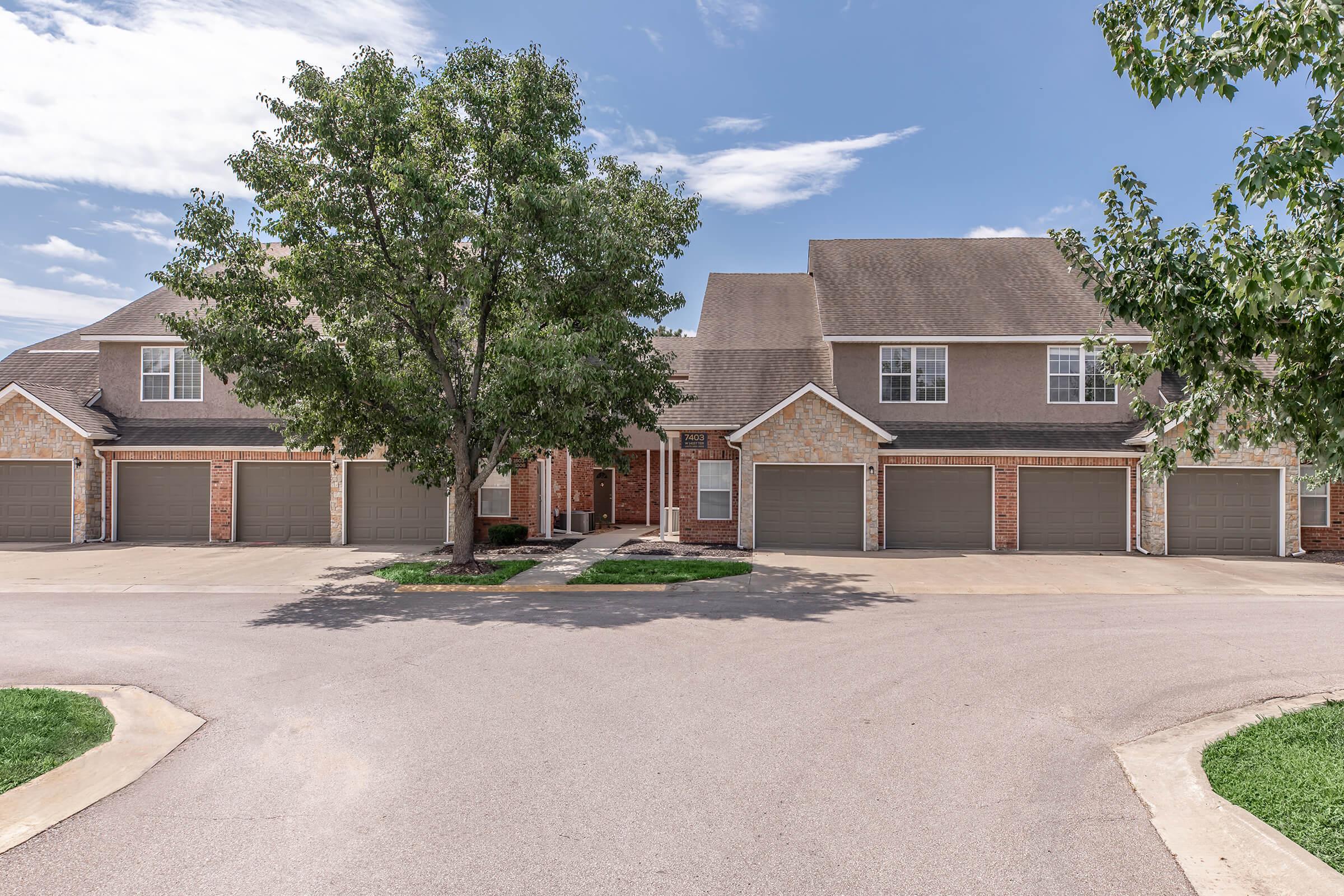 A row of brick townhouses with grey garage doors, situated in a suburban area. Lush green trees and a circular driveway in front. The sky is partly cloudy, creating a bright atmosphere.