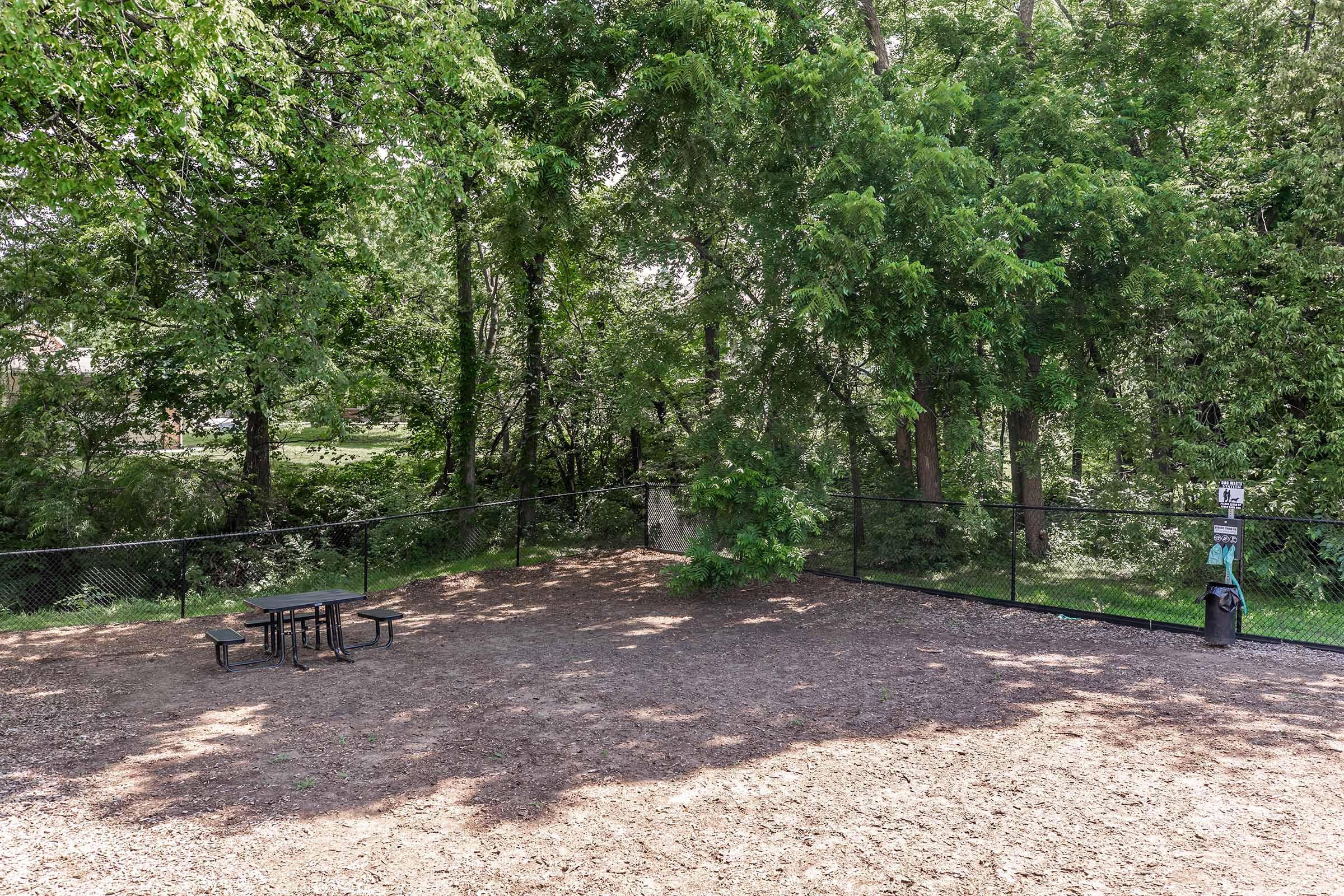 An outdoor area with a fenced space featuring a picnic table surrounded by lush green trees. The ground is covered with mulch, and there's a trash receptacle in the corner, indicating a park-like setting suitable for gatherings or relaxation.