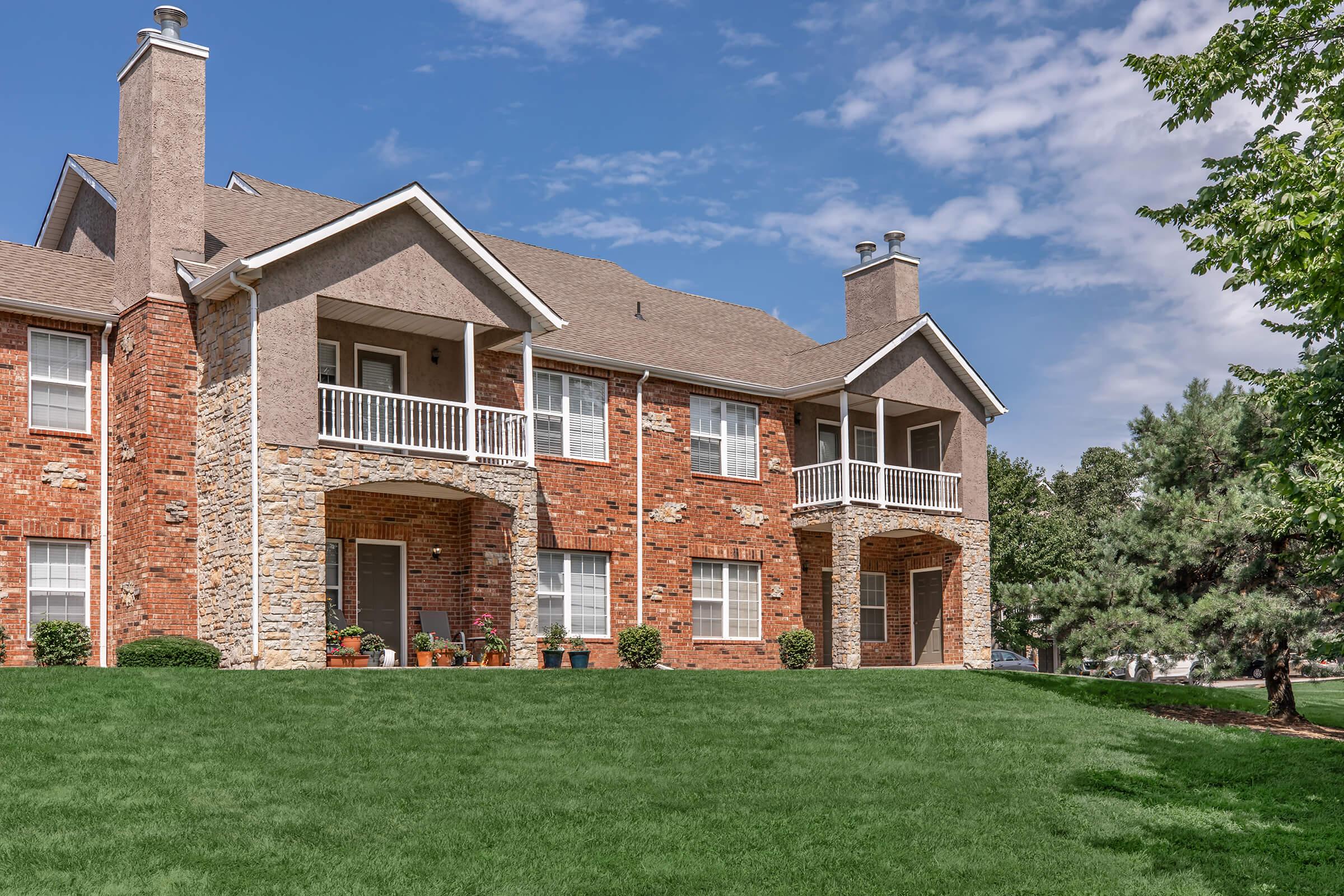 A two-story brick apartment building with stone accents, featuring four balconies with white railings. The front yard is lush green grass, and there are small bushes landscaping the area. The sky is clear with a few clouds, creating a bright and inviting atmosphere.