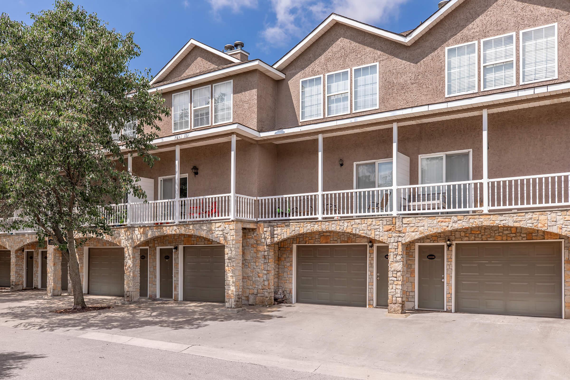 Row of townhouses with beige stucco exteriors and stone accents. Each unit has a covered balcony and a garage with a closed door. Lush green trees are present in front, and the sky is clear with a few clouds. The overall setting is suburban and inviting.