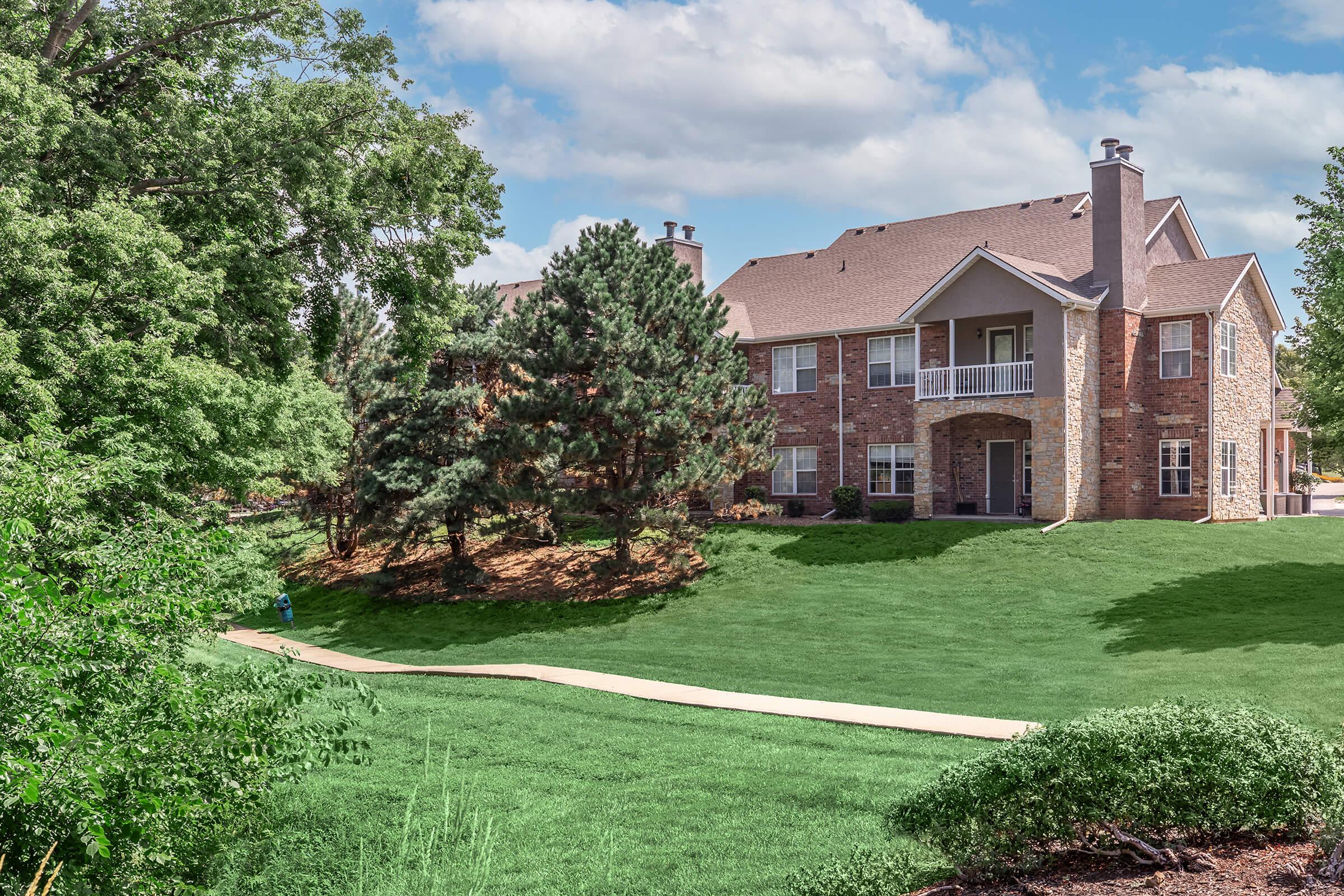 A large two-story brick apartment building surrounded by lush green grass and trees, with a clear blue sky and fluffy clouds in the background. A pathway leads to the entrance, creating a welcoming outdoor space.