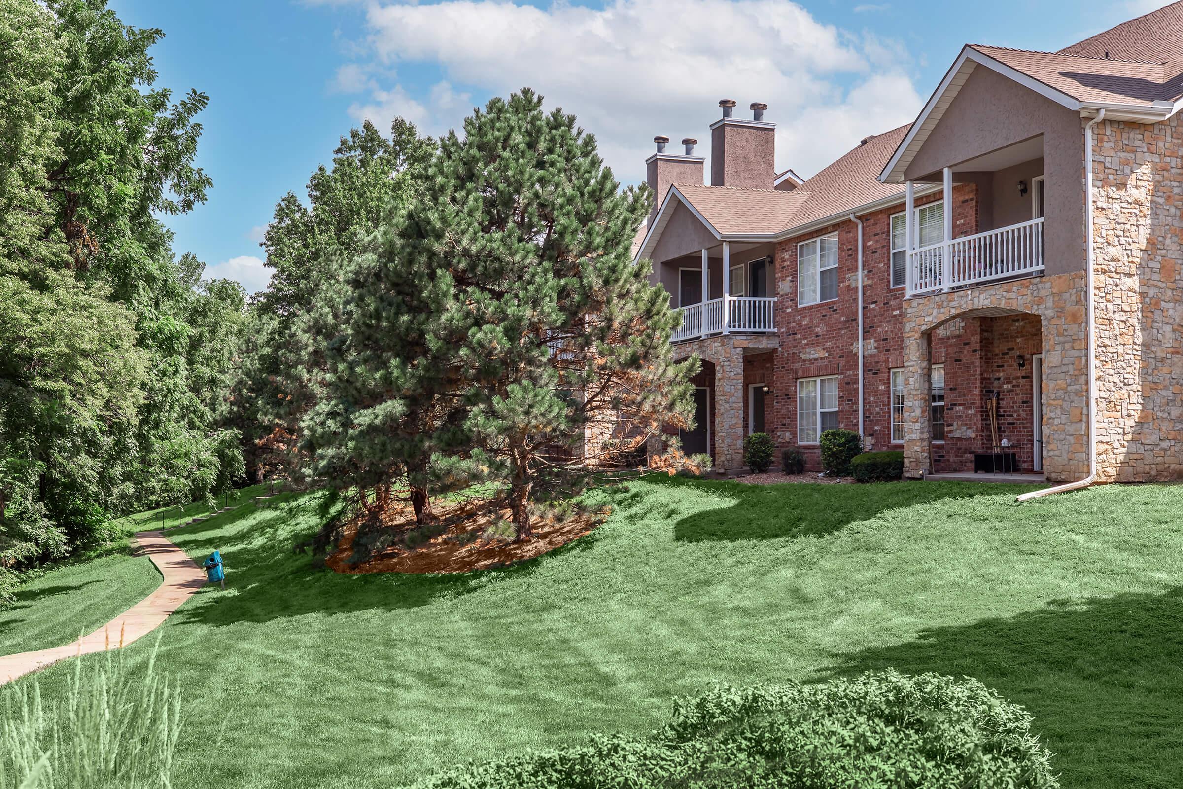 A residential building surrounded by lush green grass and trees. The building features a mix of brick and stone exterior with balconies. A pathway leads through the landscape, enhancing the serene atmosphere of the surroundings under a partly cloudy sky.