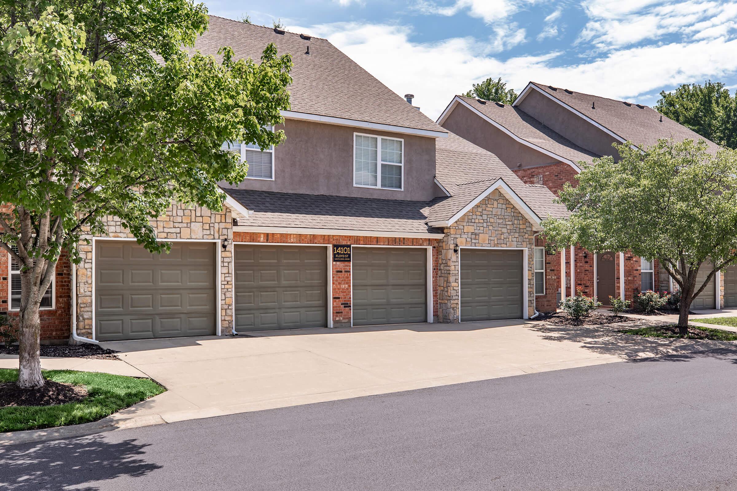 A residential street view featuring several townhouses with stone and brick facades. The focus is on a driveway with three closed garage doors, flanked by neatly trimmed shrubs and small trees. The sky is partly cloudy, enhancing the suburban atmosphere.