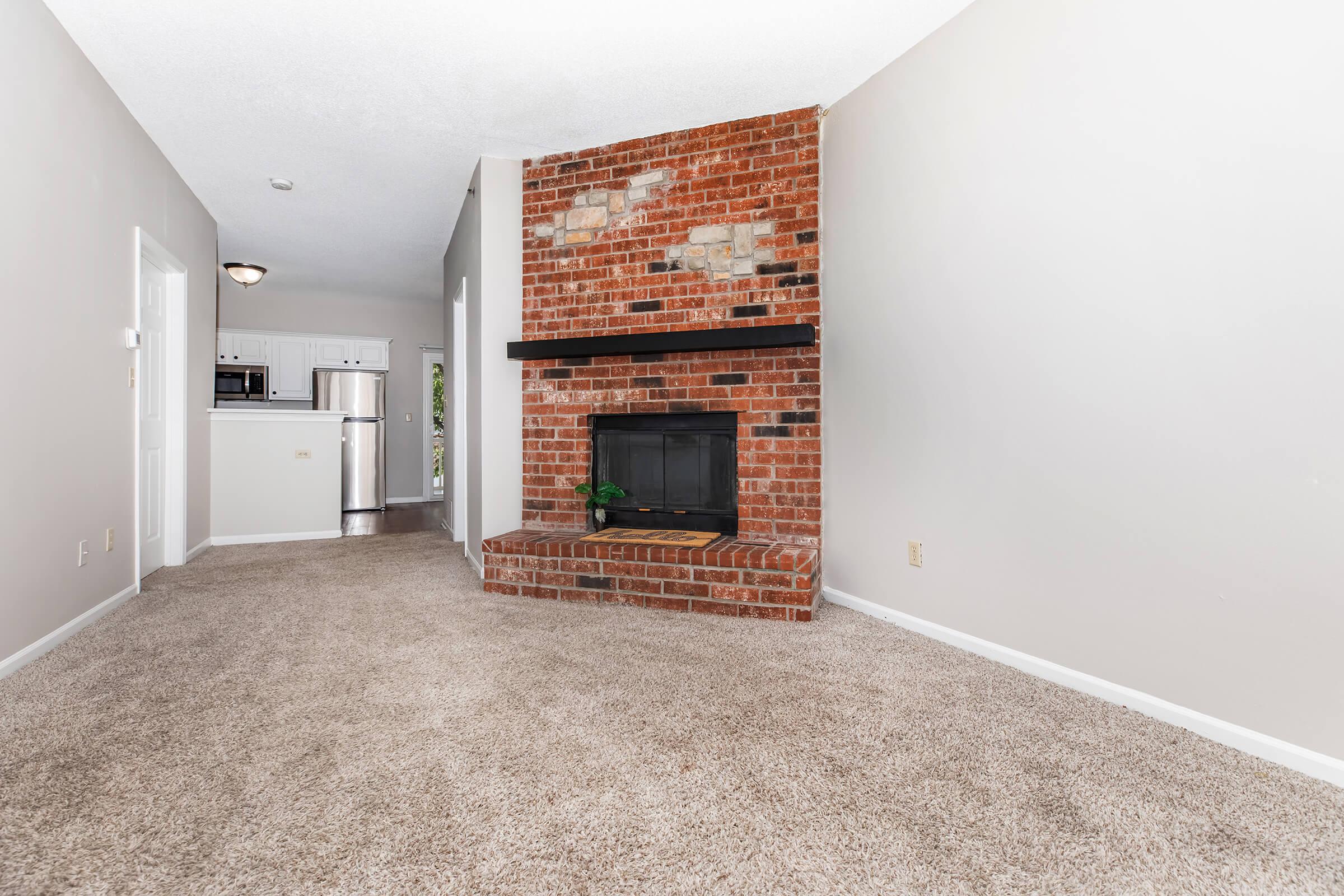 A cozy living room featuring a brick fireplace as a focal point, carpeted flooring, and light gray walls. In the background, there is a glimpse of an open kitchen with white cabinetry and stainless steel appliances, creating a warm and inviting atmosphere. Natural light fills the space.