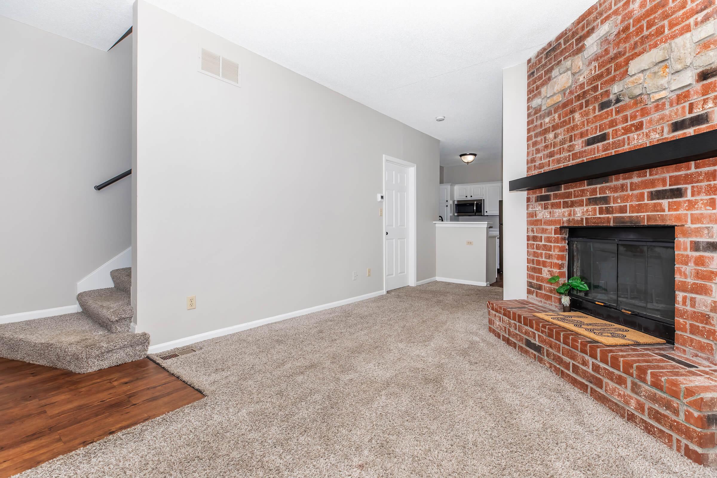 Interior view of a living room featuring light gray walls, a cozy carpeted area, a brick fireplace with a black mantle, and a staircase leading to the upper floor. The kitchen is visible in the background, with a white door and light fixtures adding to the ambiance.