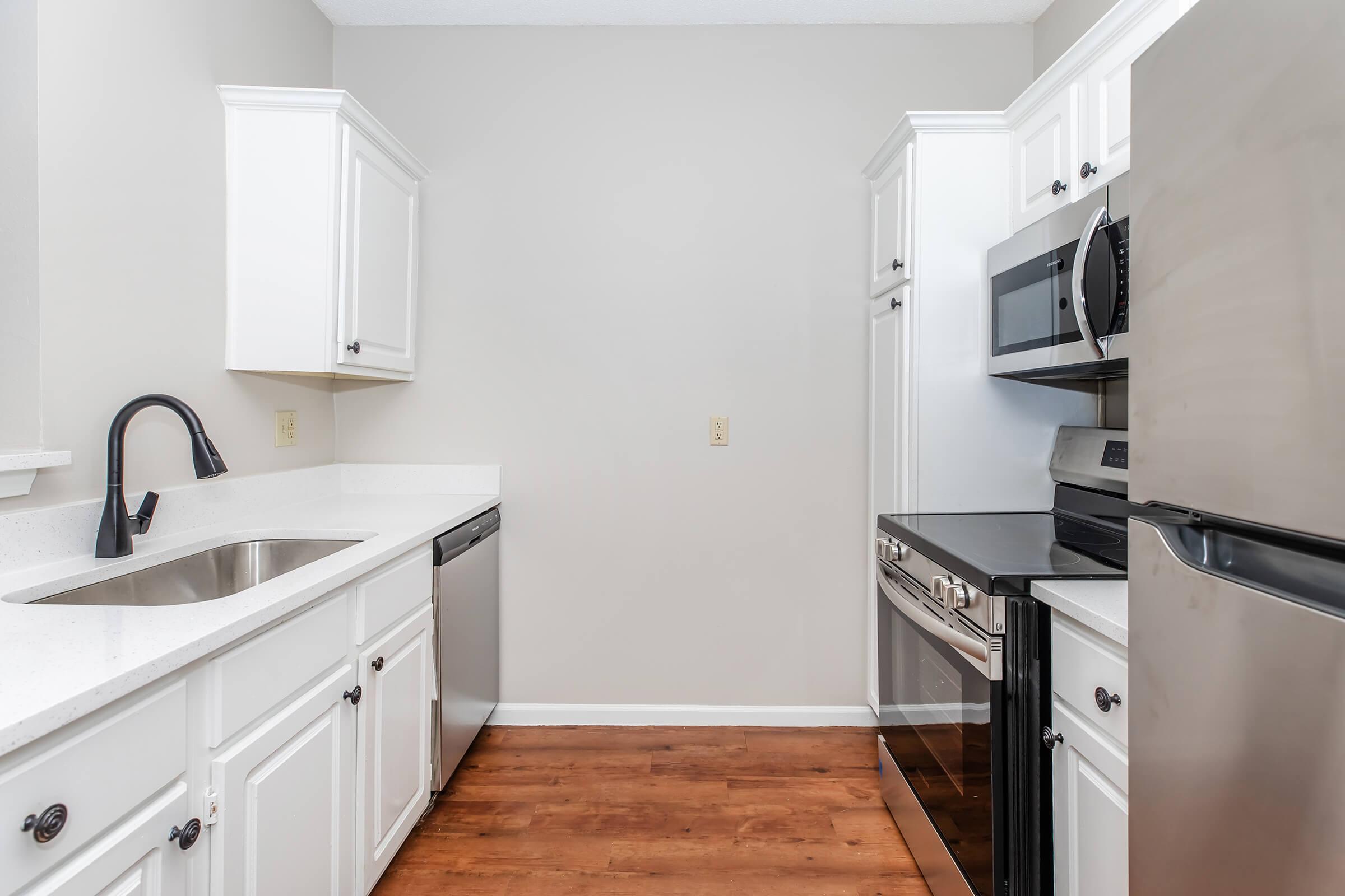 Modern kitchen featuring white cabinetry, stainless steel appliances including a microwave, oven, and dishwasher, along with a sleek sink. The countertop is light-colored and the flooring is a warm wood tone, creating a bright and spacious ambiance.