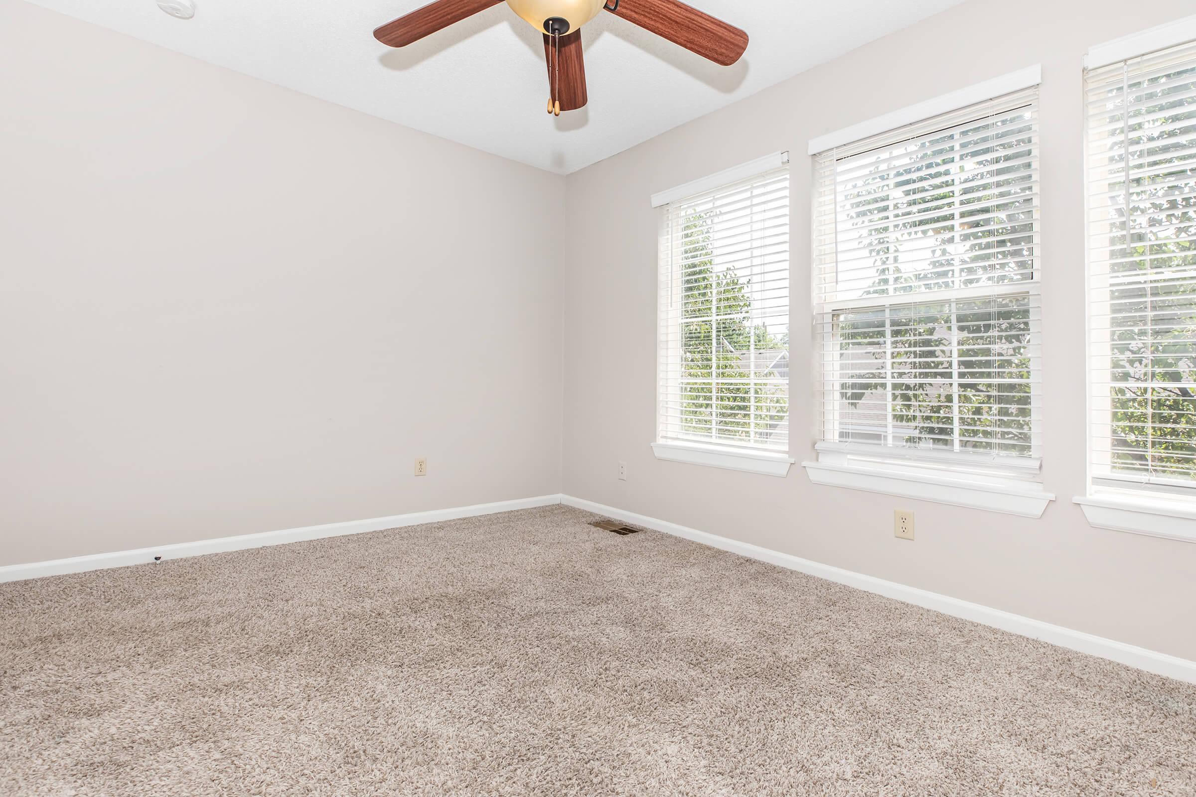 A spacious, empty room featuring light beige walls and a carpeted floor. It has two large windows with white blinds, allowing natural light to flood in. A ceiling fan with wooden blades hangs from the ceiling. The room is well-lit and invites a sense of openness and potential for decoration.