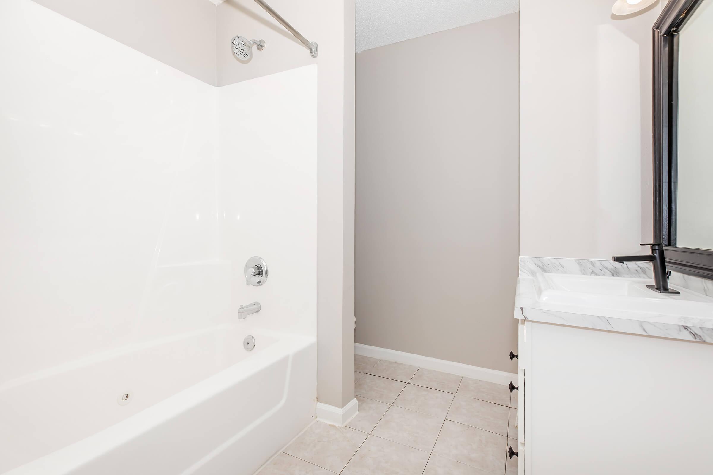 A clean bathroom featuring a white bathtub with a shower fixture, beige walls, and a spacious vanity with a black mirror. The floor is tiled, and the overall design is bright and contemporary, emphasizing a minimalist aesthetic.