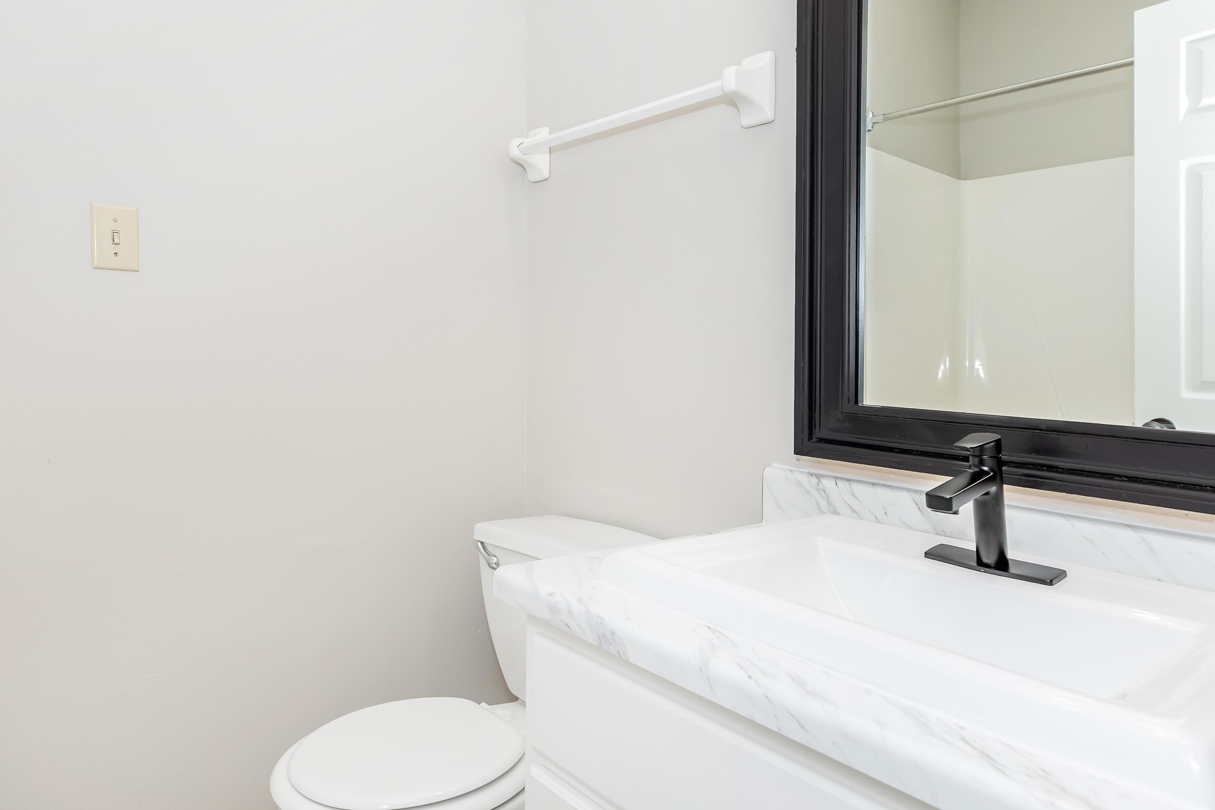 A modern bathroom featuring a sleek white sink with a black faucet, a white toilet, and a frameless mirror. The walls are painted a light gray, and there is a towel bar mounted on the wall. The space is clean and minimalistic, with natural light illuminating the area.