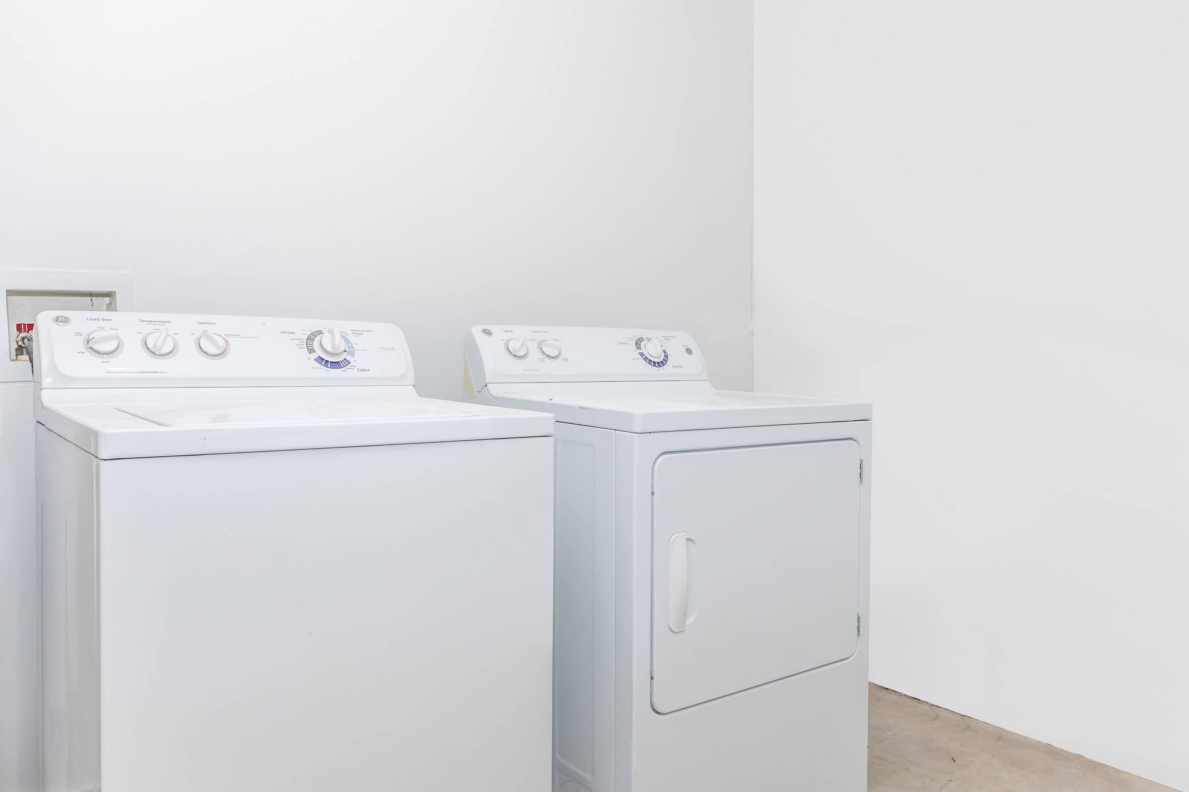A white washing machine and a white dryer are side by side against a plain white wall, with a light-colored floor below them. The appliances have simple controls on the front.