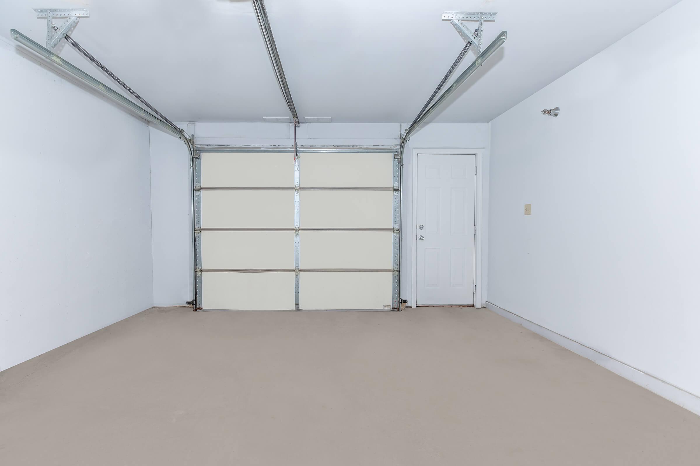 Interior of a clean, empty garage featuring white walls, a beige floor, and a closed garage door. A side door is visible on the right. The space is well-lit, with no other furnishings or clutter.