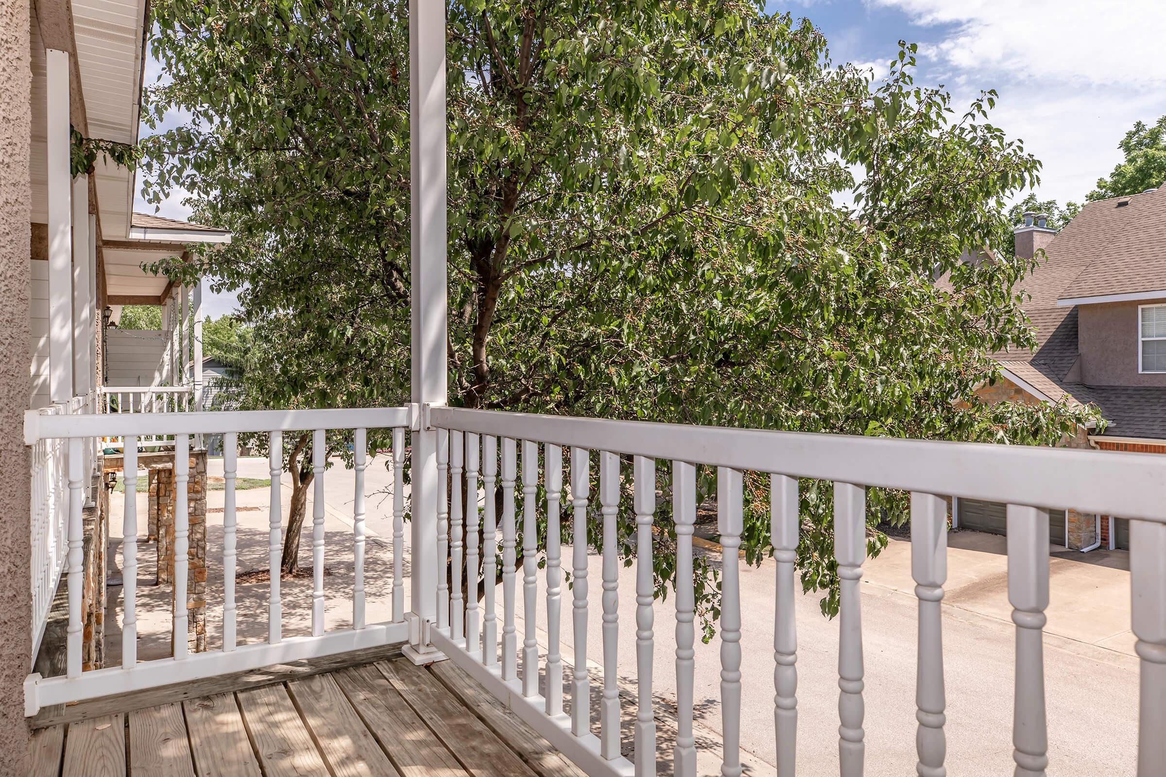 A wooden balcony with white railings, overlooking a tree and a street below. The scene is bright with blue skies and fluffy clouds, creating a peaceful outdoor atmosphere. The nearby buildings are partially visible, adding to the residential setting.